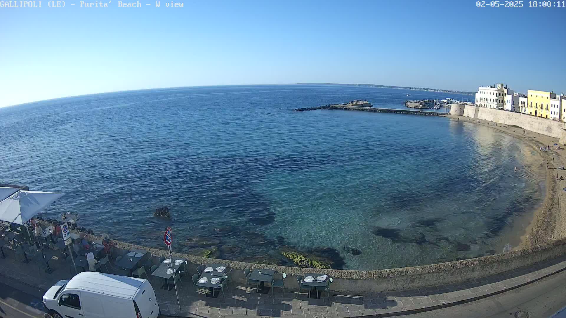 A clear, sunny day reveals a tranquil bay with calm, clear turquoise water lapping a sandy beach, lined by a stone wall and outdoor restaurant seating, with a small white van parked nearby.