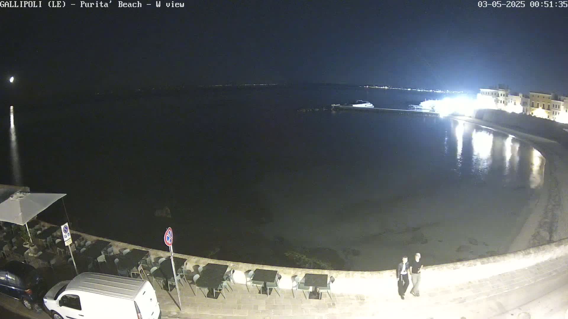 A nighttime view of a calm, dark body of water next to a shoreline with outdoor seating and two people standing near a wall.