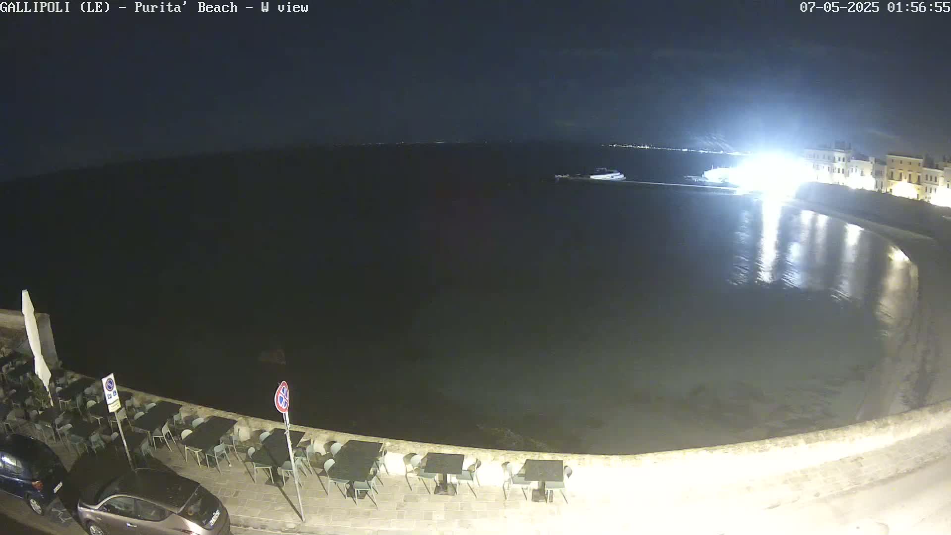 A nighttime view of a calm, dark body of water next to a stone wall with outdoor seating, and a few cars parked nearby.