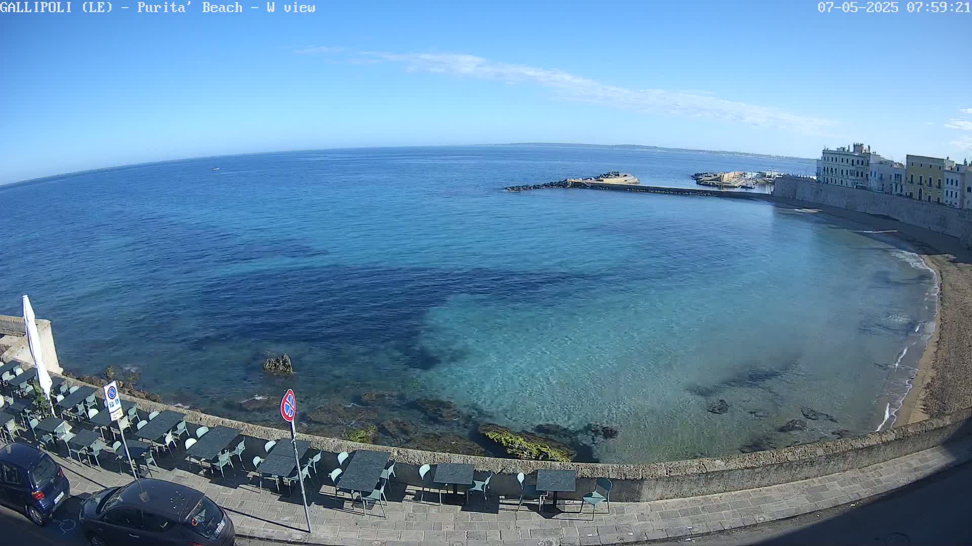 A clear, sunny day reveals a picturesque coastal scene with a calm, turquoise sea lapping against a rocky shore lined with outdoor seating.