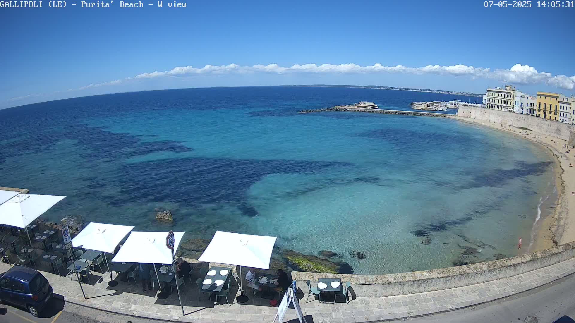 A partly sunny day shows a beach with clear, shallow turquoise water, a stone wall with buildings and a few people sitting at outdoor tables under umbrellas.