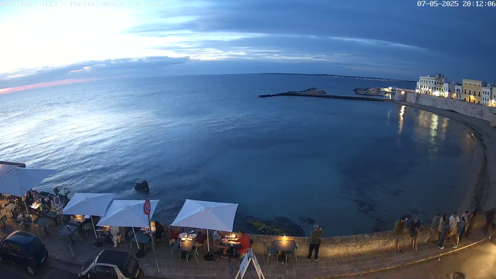 An outdoor waterfront restaurant with white umbrellas and patrons sits along a calm, dark-blue sea at twilight under a mostly clear sky.