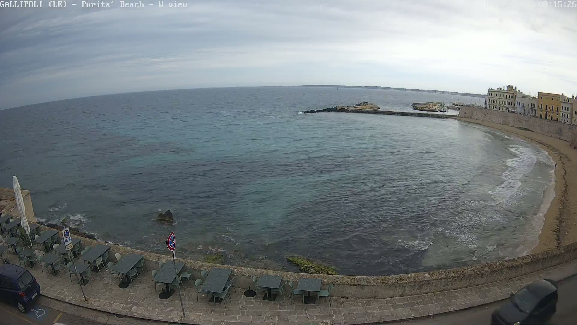 A partly cloudy day overlooks a calm bay with a sandy beach and stone buildings, featuring outdoor seating along a seaside wall.