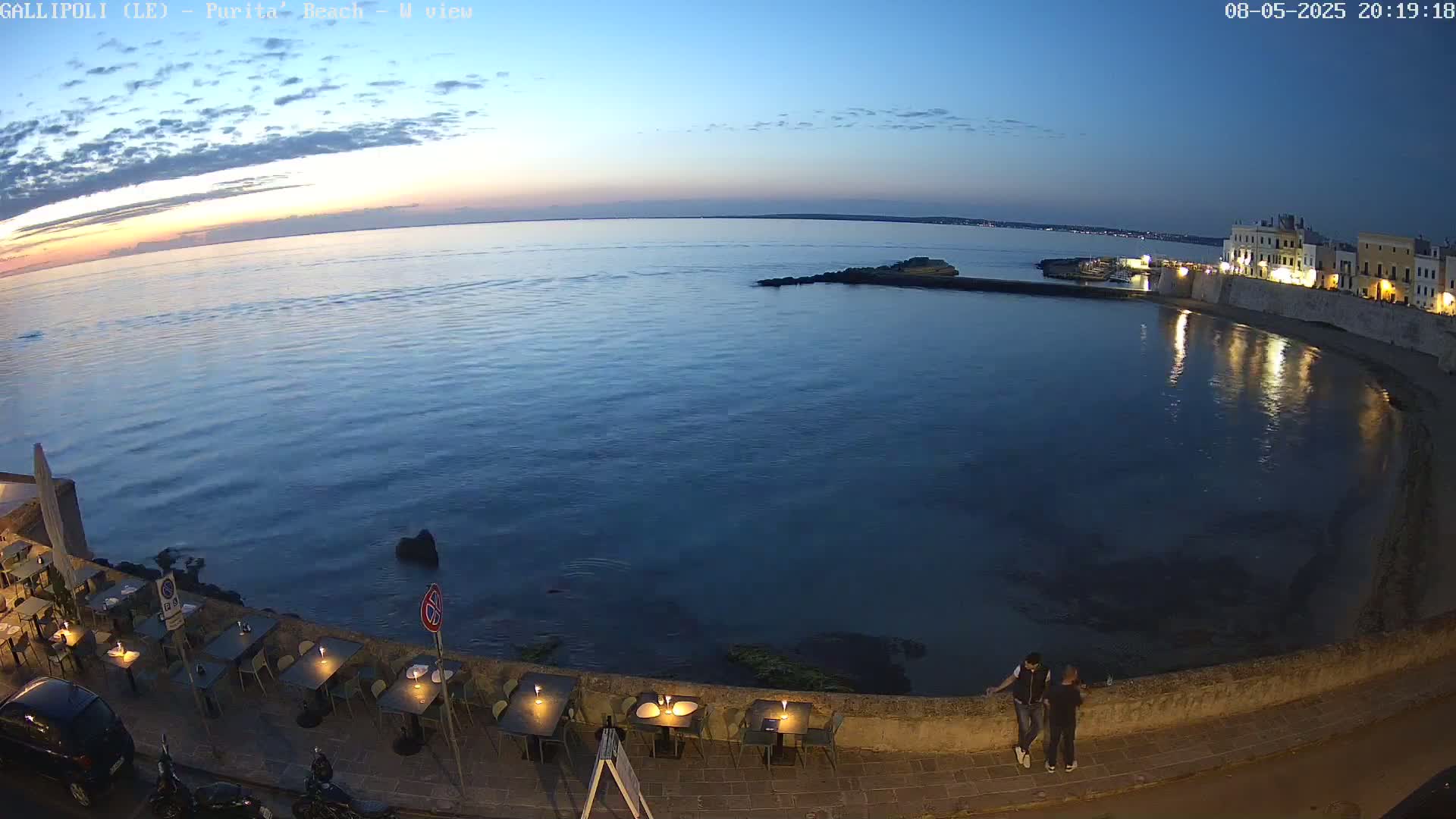 A calm, dark-blue sea at twilight is viewed from an elevated position, showing a seaside restaurant with outdoor seating and two people standing near a low wall overlooking the water.
