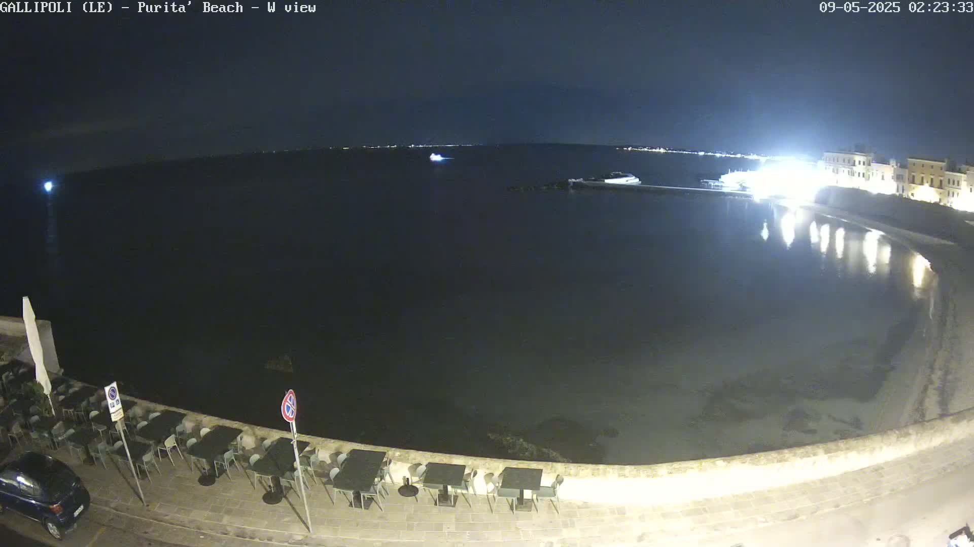 A nighttime view of a calm, dark body of water next to a waterfront area with outdoor seating, under a clear, dark sky.