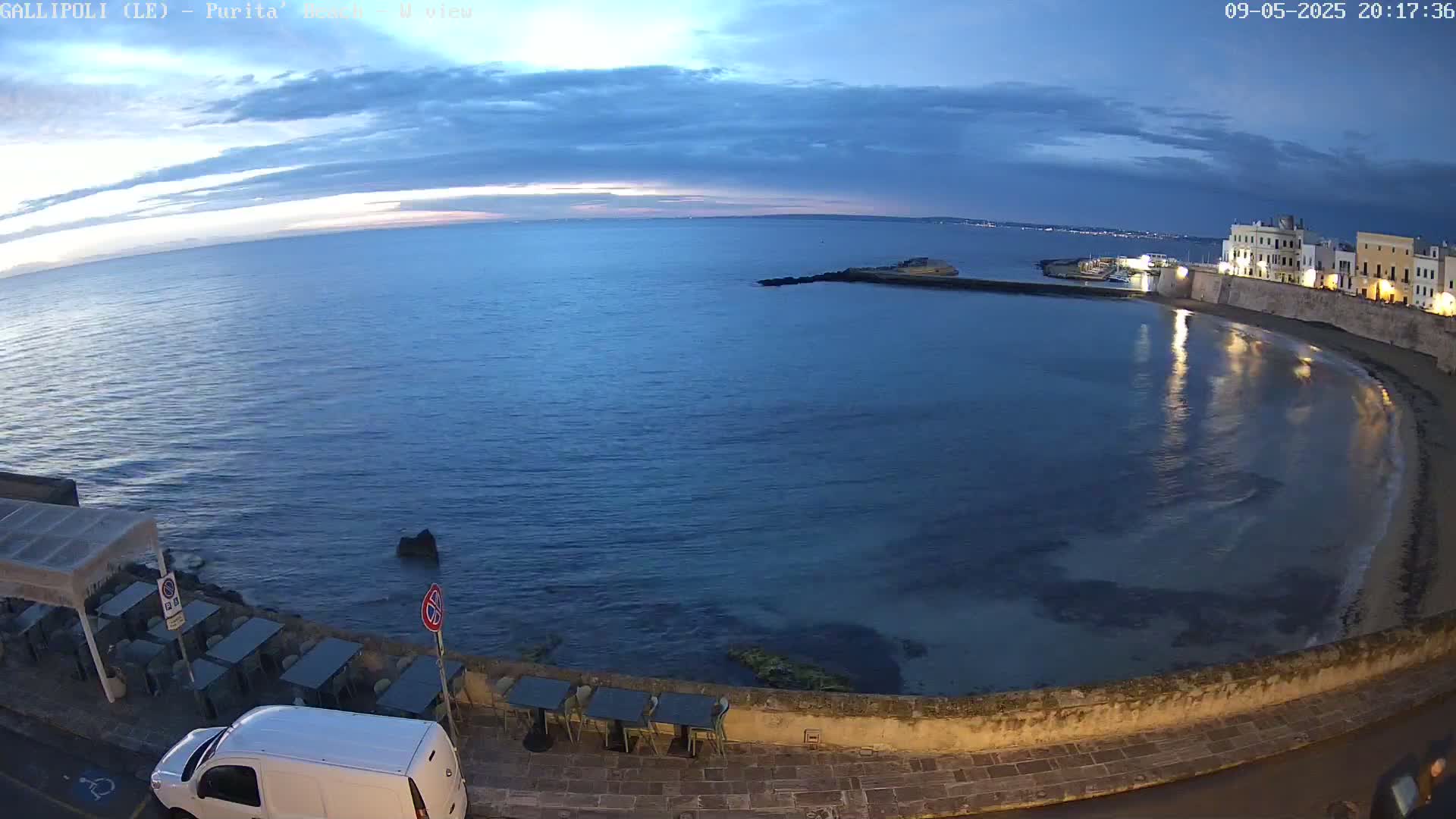 A coastal town at dusk, with calm dark-blue water lapping a gently curving shoreline under a mostly clear sky.