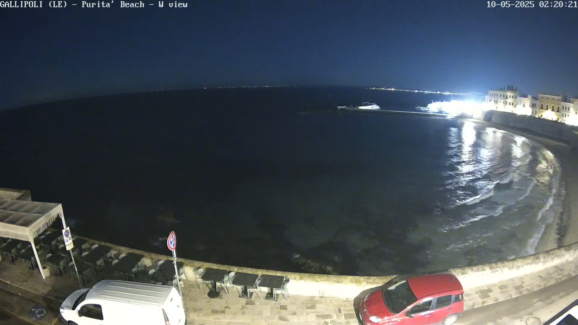 A nighttime view of a calm, dark sea lapping gently at a stone wall beside a paved area with outdoor seating and parked vehicles.