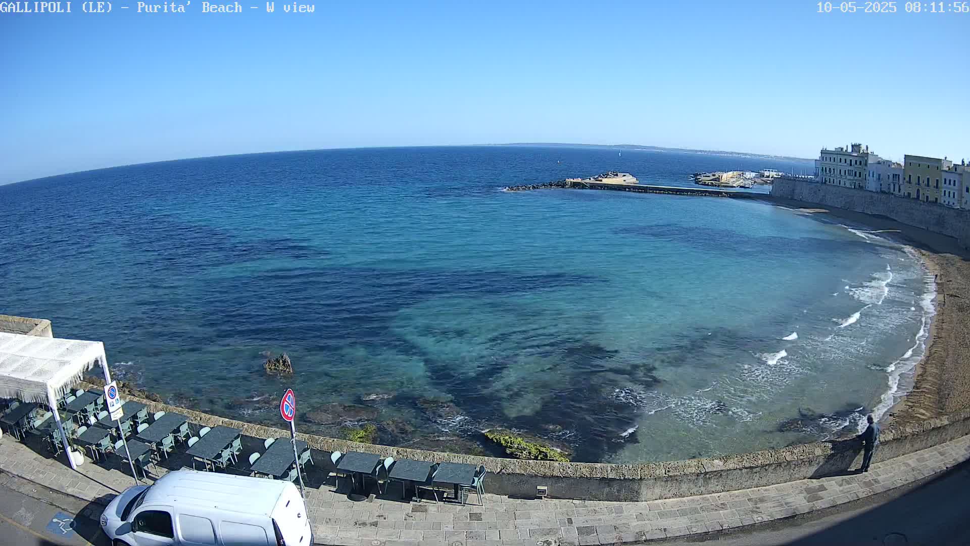 A clear, sunny day reveals a calm, turquoise sea lapping a sandy beach next to a stone wall and buildings; outdoor cafe tables and chairs are set up near the water's edge.