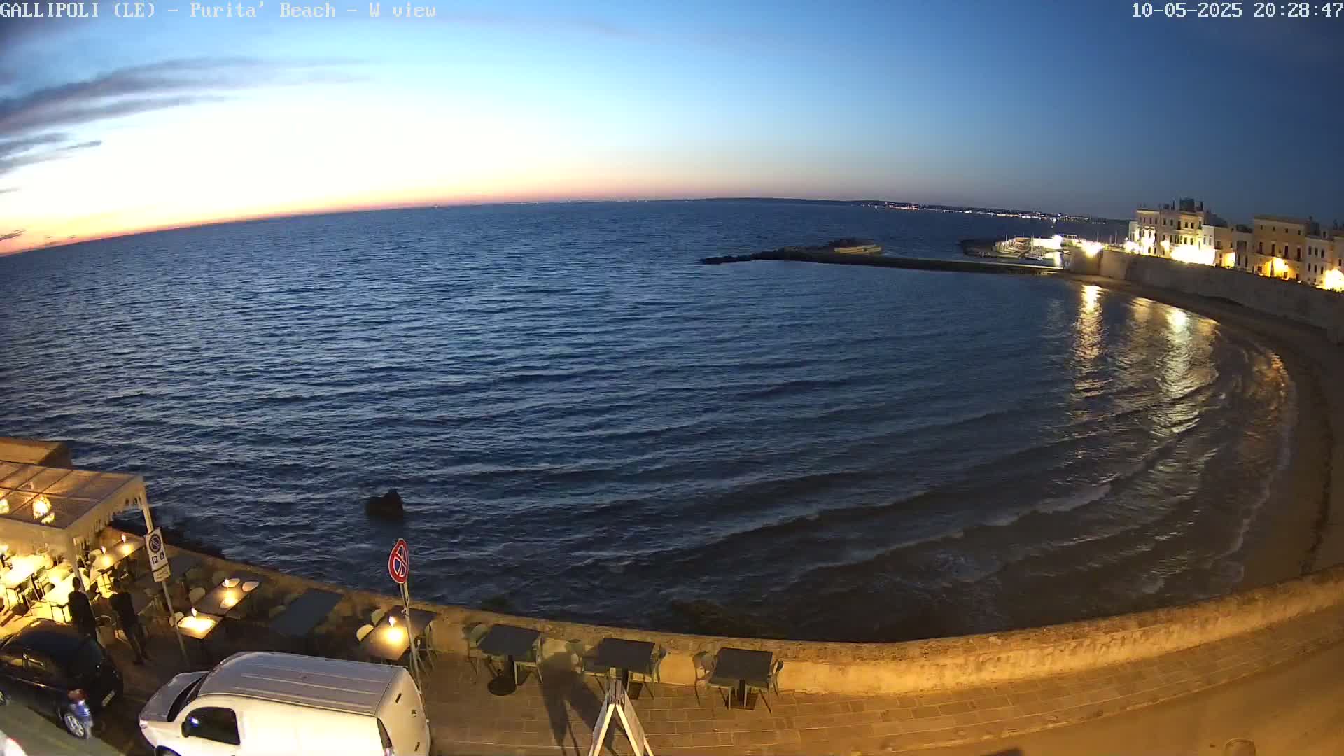 A seaside town at twilight, with calm waves lapping a curved shore lined by a wall and outdoor seating areas.