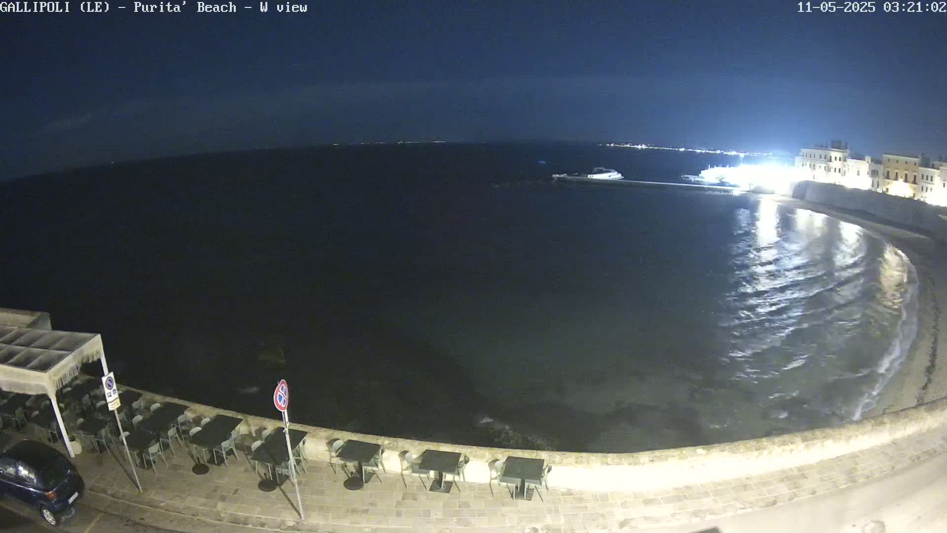 A nighttime view shows a calm sea lapping a stone embankment next to an outdoor cafe with empty tables and chairs, and buildings lit up in the background.