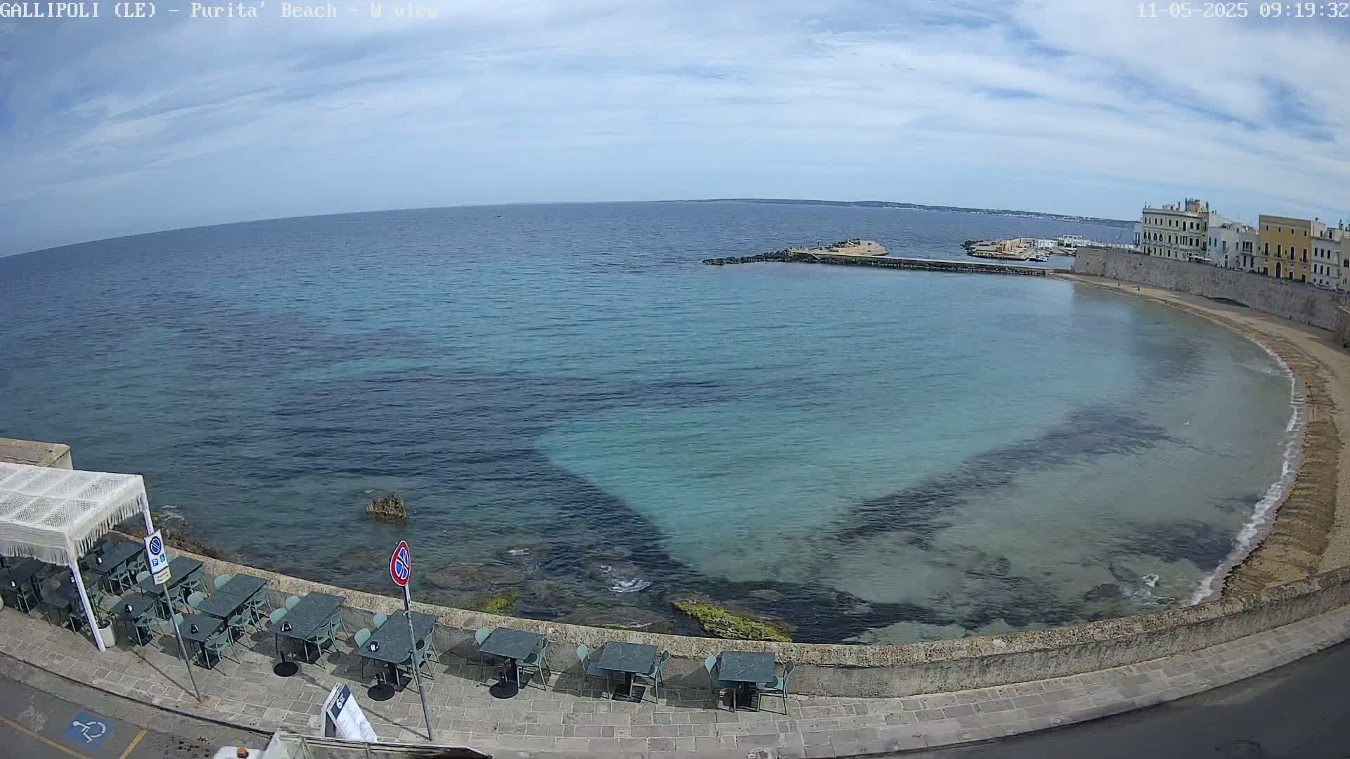 A clear, sunny day overlooks a calm, turquoise bay with a sandy beach curving around a stone wall lined with outdoor seating.