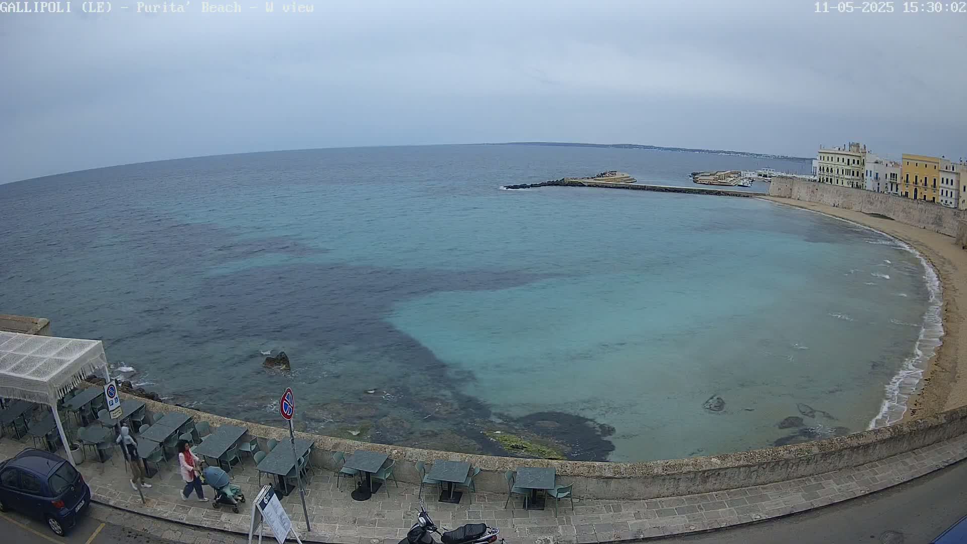 A partially cloudy day shows a tranquil, turquoise bay curving around a stone wall lined with outdoor seating, near a town with pale buildings.