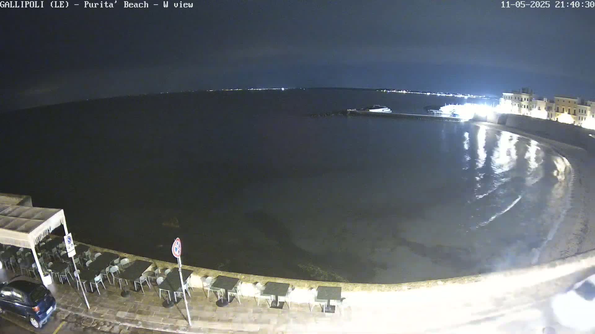 A nighttime, seaside view shows a calm body of water reflecting lights from a building and a line of empty tables and chairs along a paved waterfront.