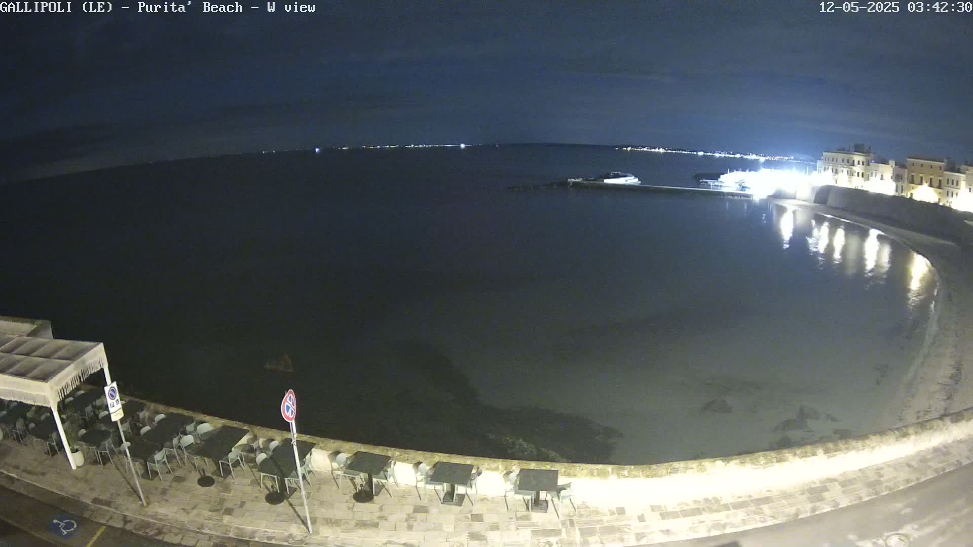 A nighttime view of a calm, dark-colored body of water next to a shoreline with outdoor seating, under a mostly clear sky.