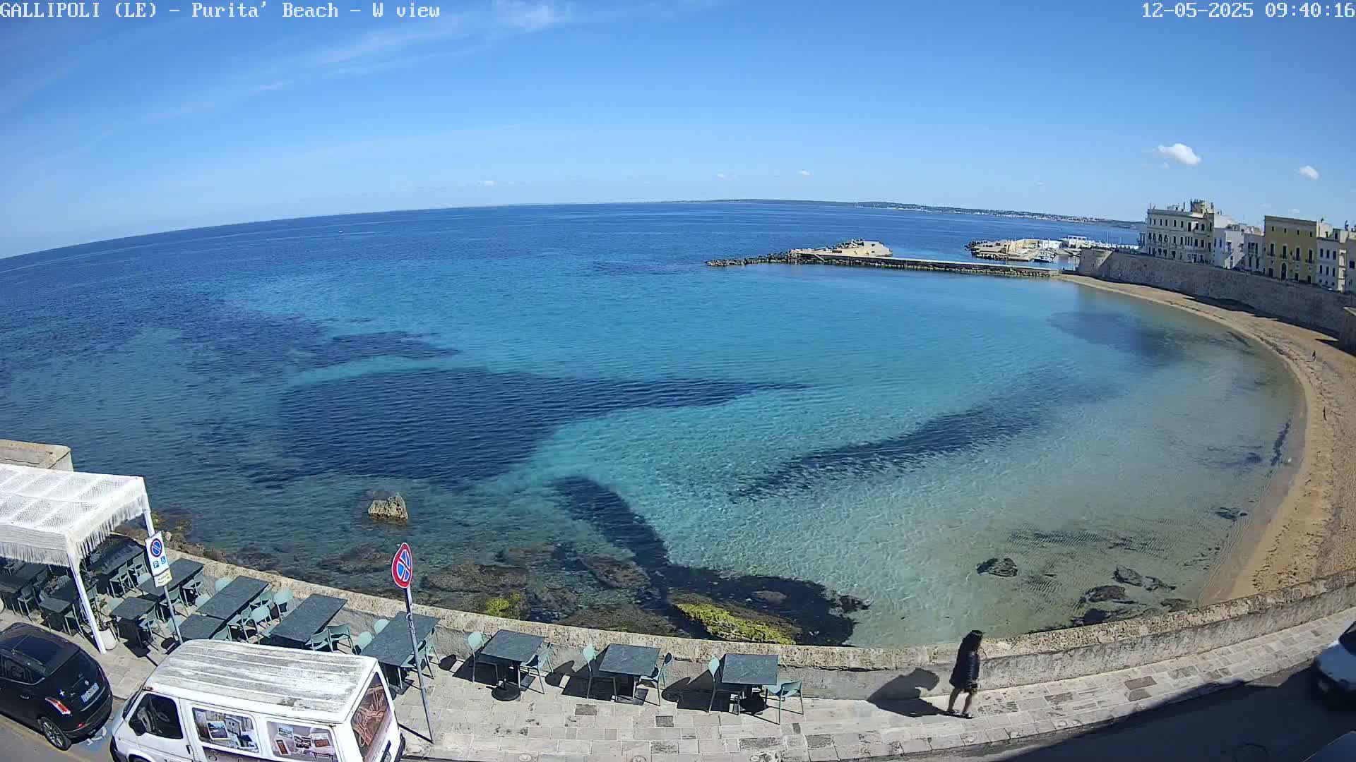 A clear, sunny day reveals a secluded, curved beach with turquoise water lapping at a stone wall, where outdoor tables and chairs are set up alongside parked vehicles and a lone person walks by.