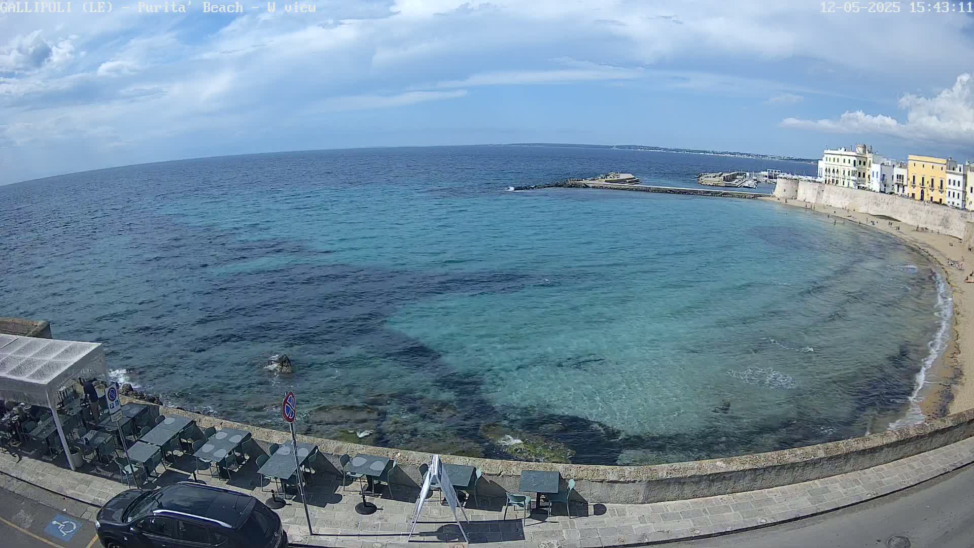 A partly sunny day shows a clear, teal-colored ocean curving around a sandy beach next to a stone wall lined with outdoor seating and a parked car.