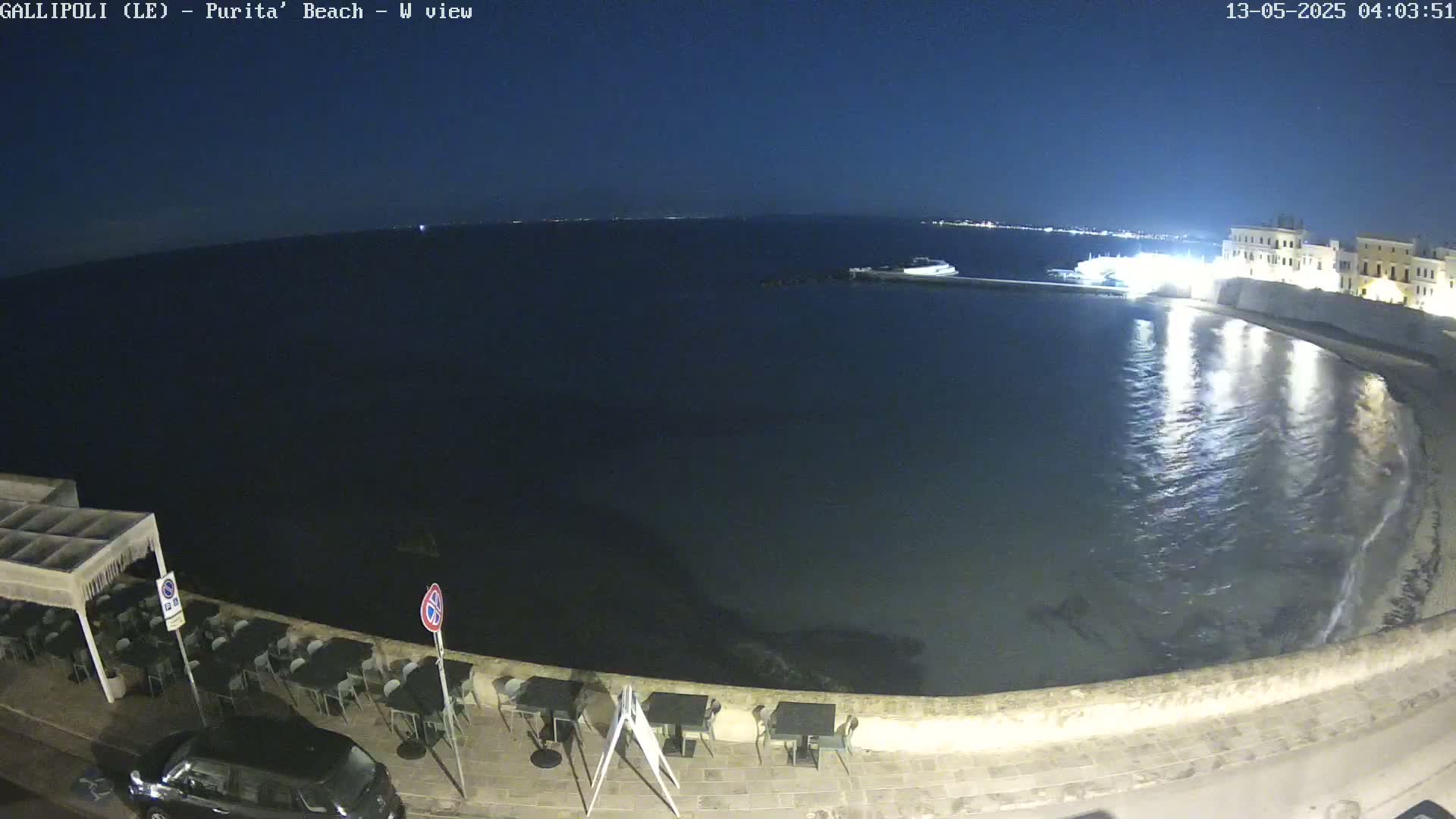A dark, calm sea laps a shoreline at night, illuminated by lights reflecting on the water from buildings and a nearby town.