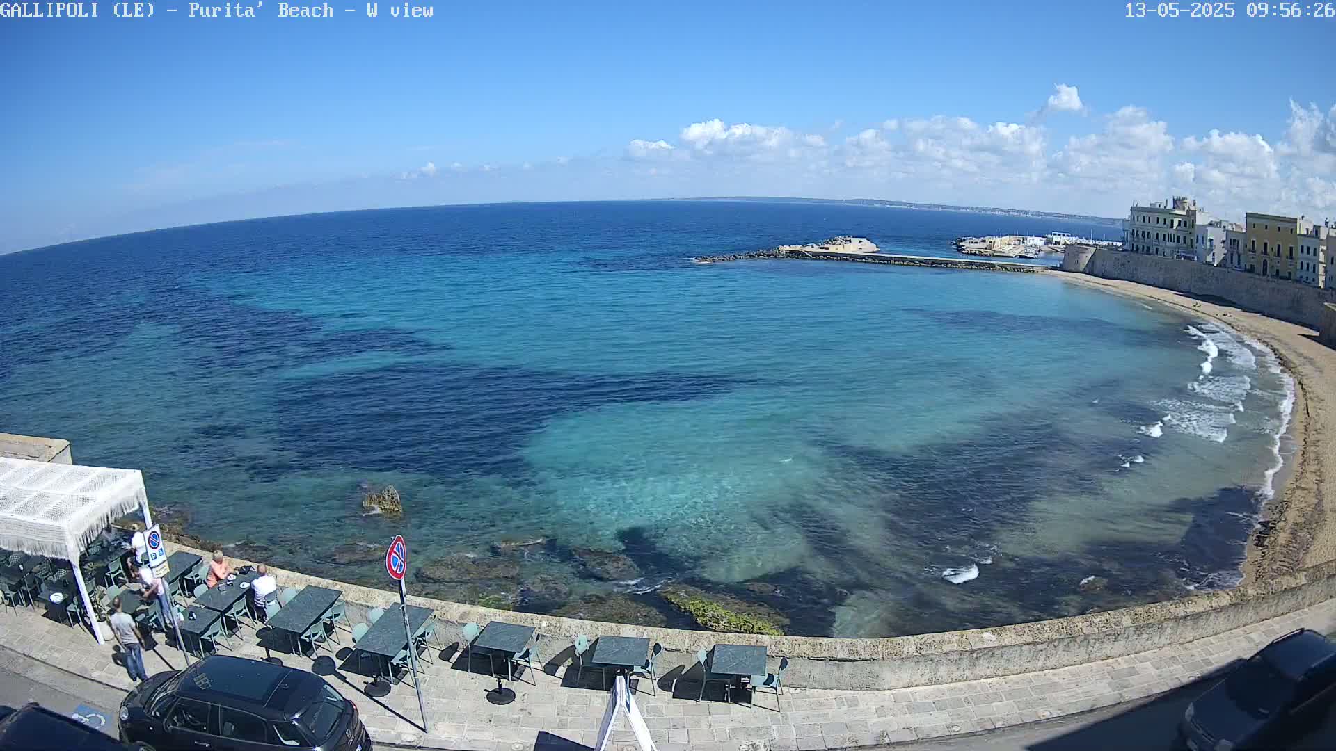 A mostly sunny day shows a clear, turquoise ocean bay with a sandy beach curving to the right, next to a stone wall and several outdoor cafe tables with people seated.