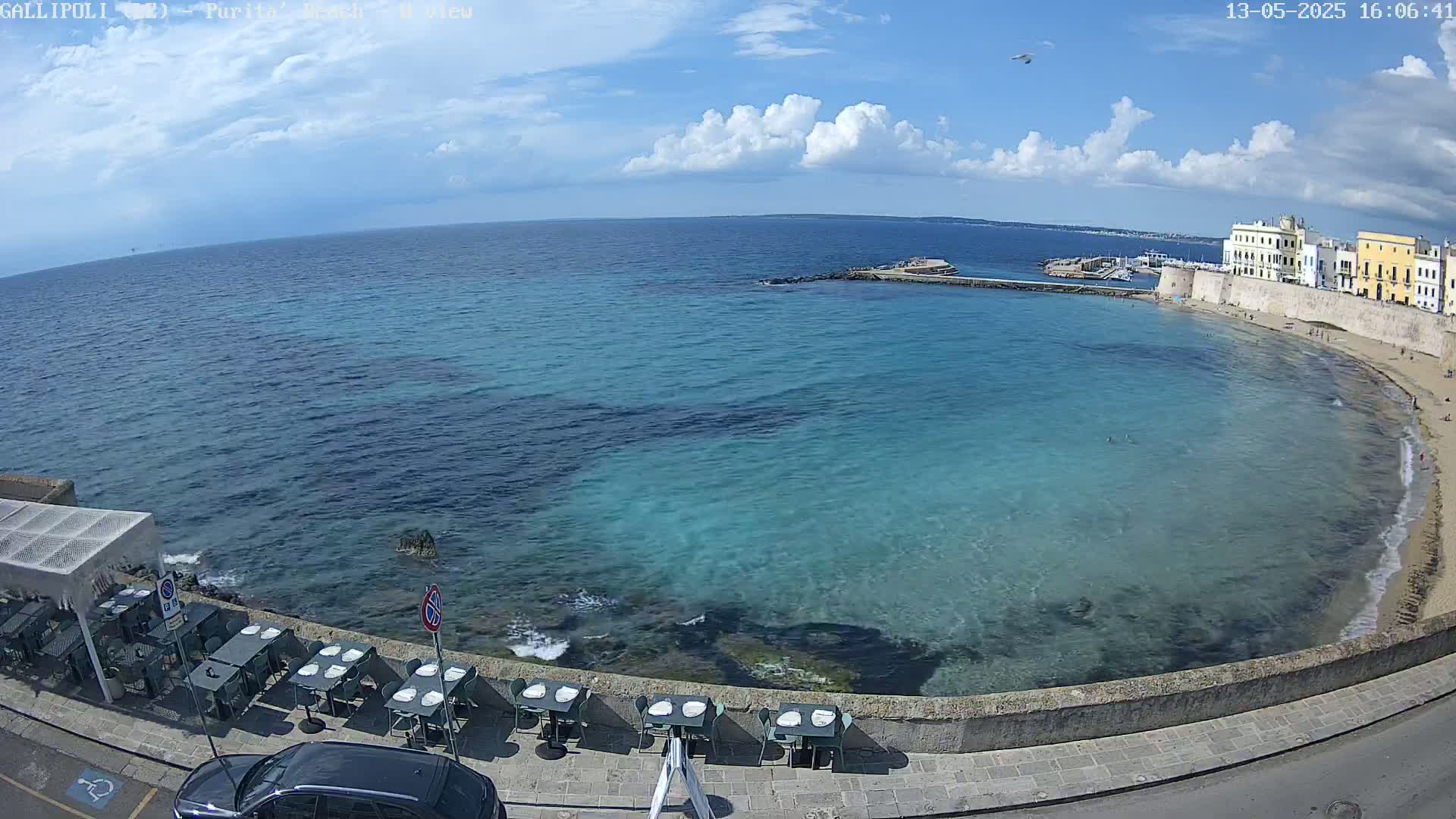 A partly sunny day shows a clear, turquoise bay with a sandy beach and a seaside restaurant with tables and chairs set up along a stone wall, next to a town with buildings along the water.