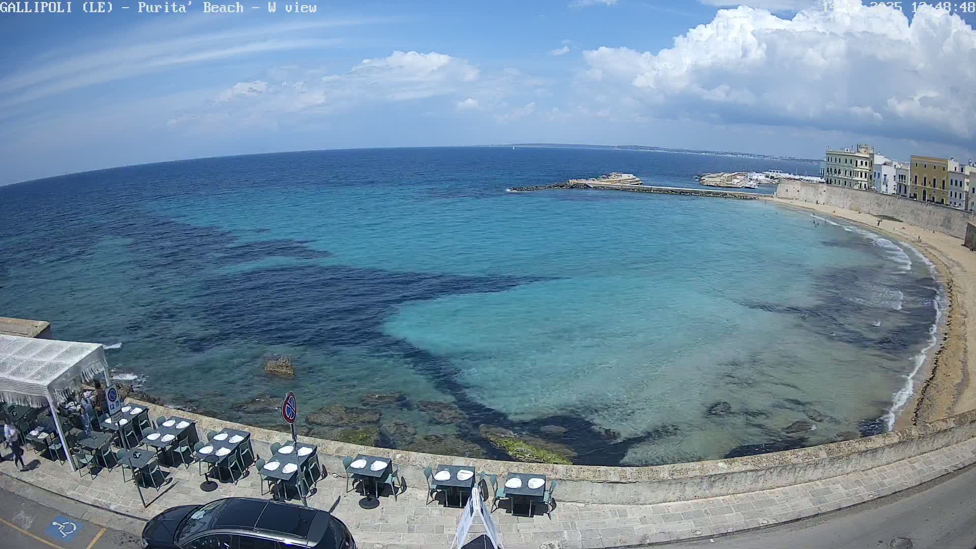 A partly sunny day shows a clear, turquoise bay with a sandy beach, lined with outdoor seating at a restaurant next to a stone wall and buildings.