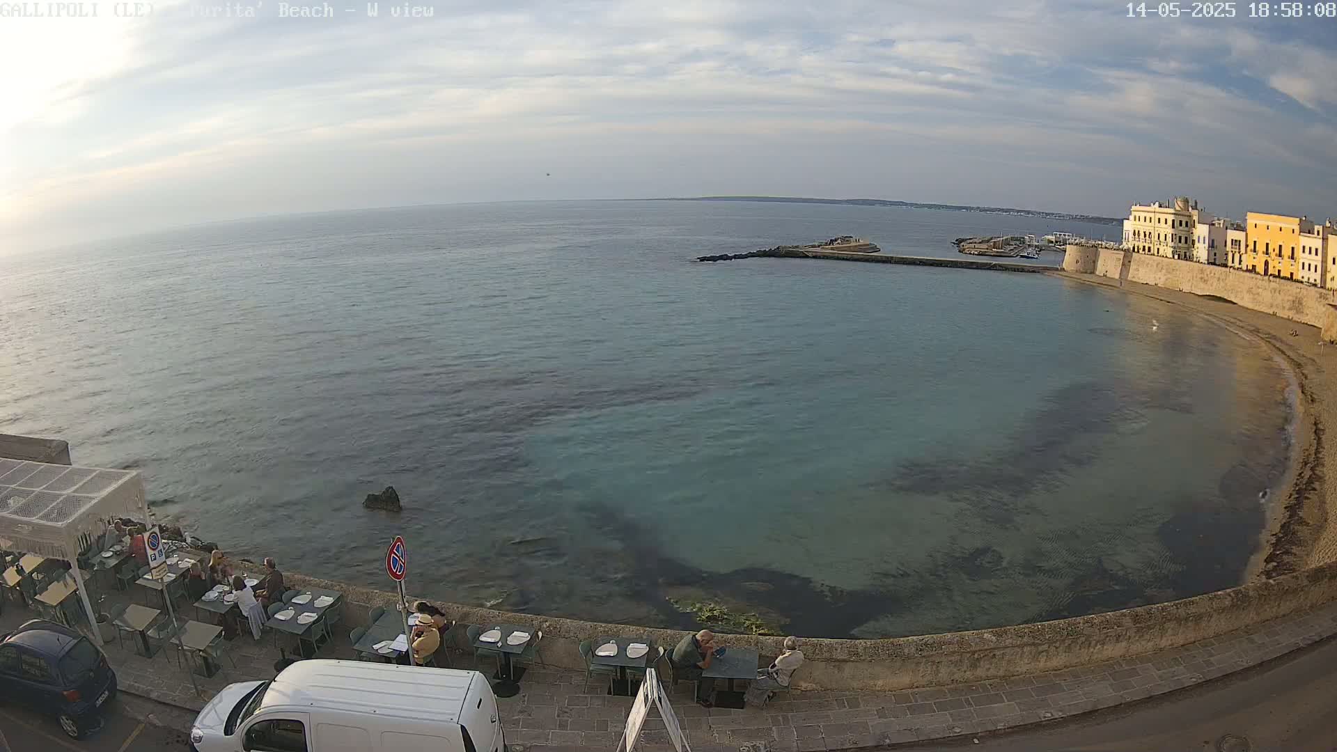 A seaside restaurant with patrons sits along a calm, clear, teal-colored bay next to a stone city wall under a partly cloudy sky.
