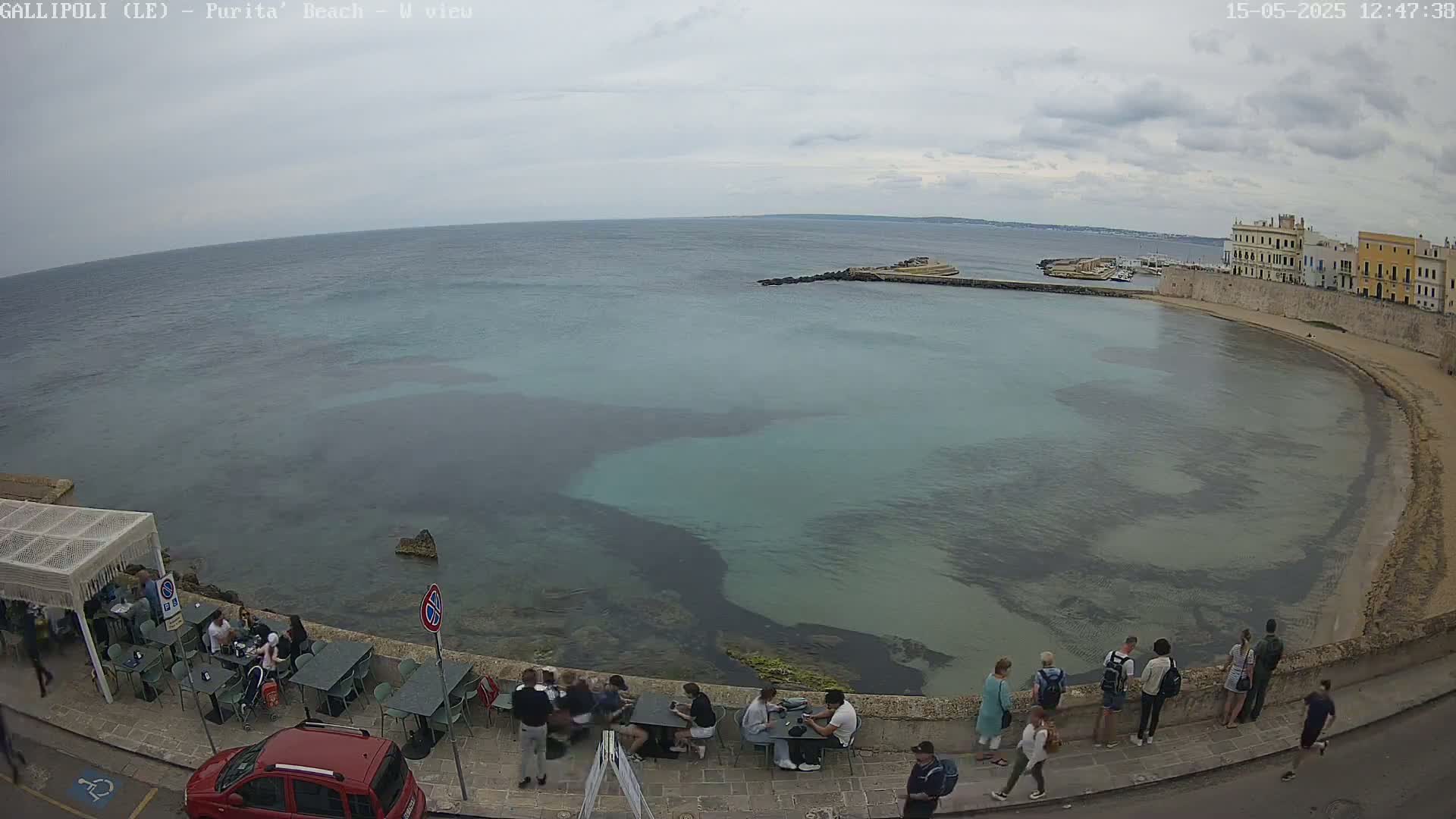 An overcast day reveals a tranquil bay with clear, shallow turquoise water, a sandy beach, and people sitting at outdoor cafes along a stone wall next to the water.
