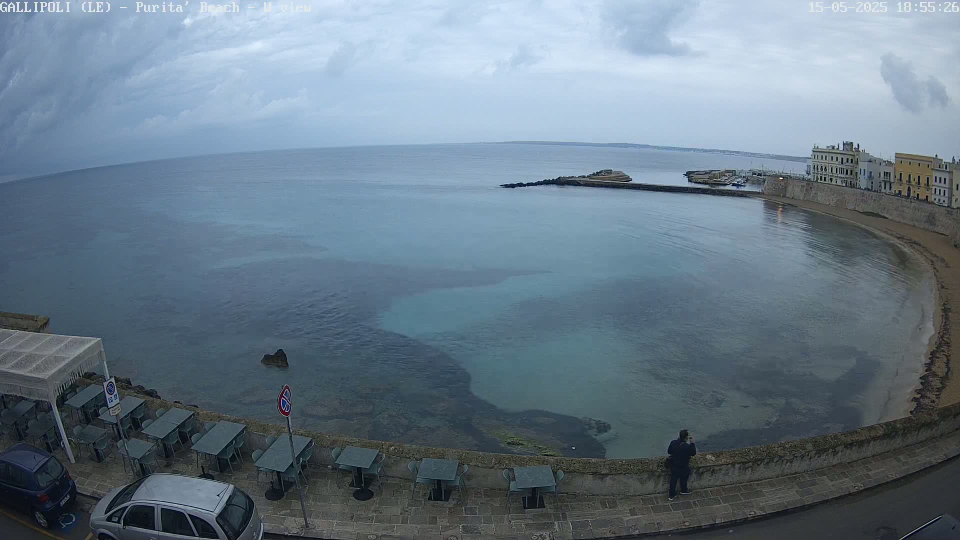 A calm, overcast day reveals a curved bay with clear, shallow teal water, a stone wall lined with outdoor seating, and a person standing by the water's edge.