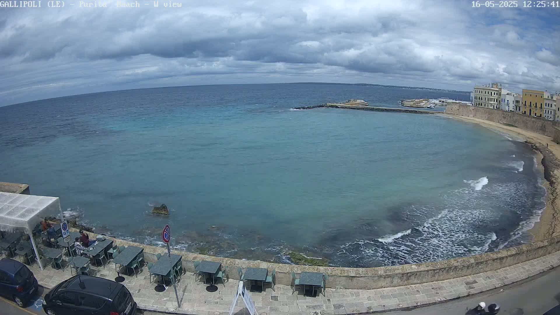 A partly cloudy day overlooks a calm, teal-colored ocean bay with a sandy beach, a stone wall, and outdoor seating at a beachfront cafe.