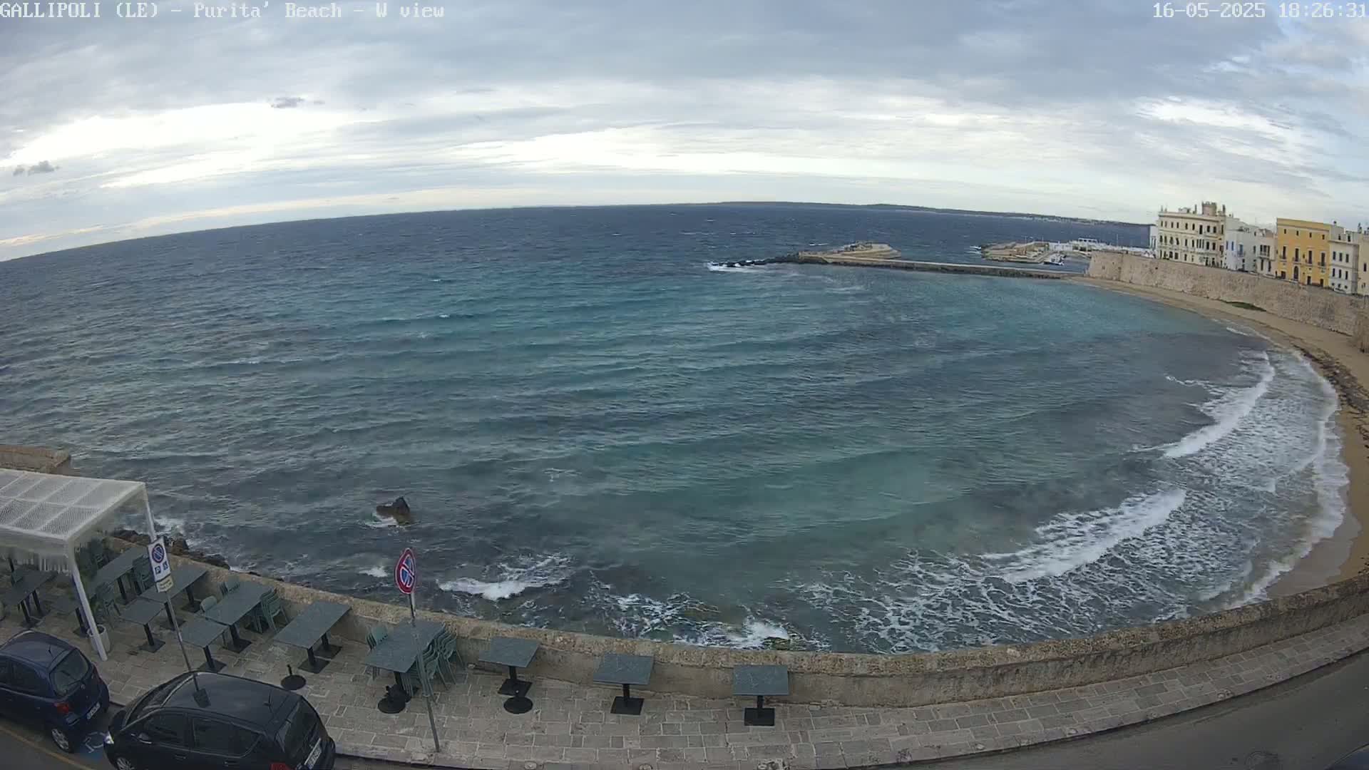 A partly cloudy day shows a coastal scene with a calm sea, small waves breaking on a sandy beach, and a row of outdoor tables near a stone wall next to parked cars.