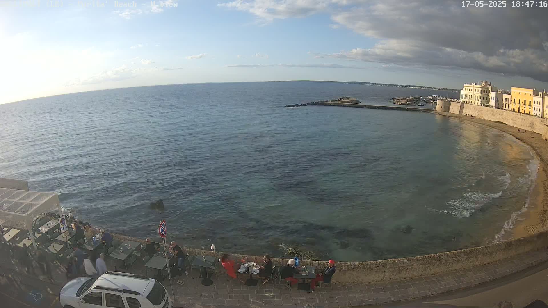 A partly cloudy day overlooks a calm sea with small waves lapping a sandy beach next to a stone wall and buildings, where several people are seated at outdoor tables at a waterfront restaurant.