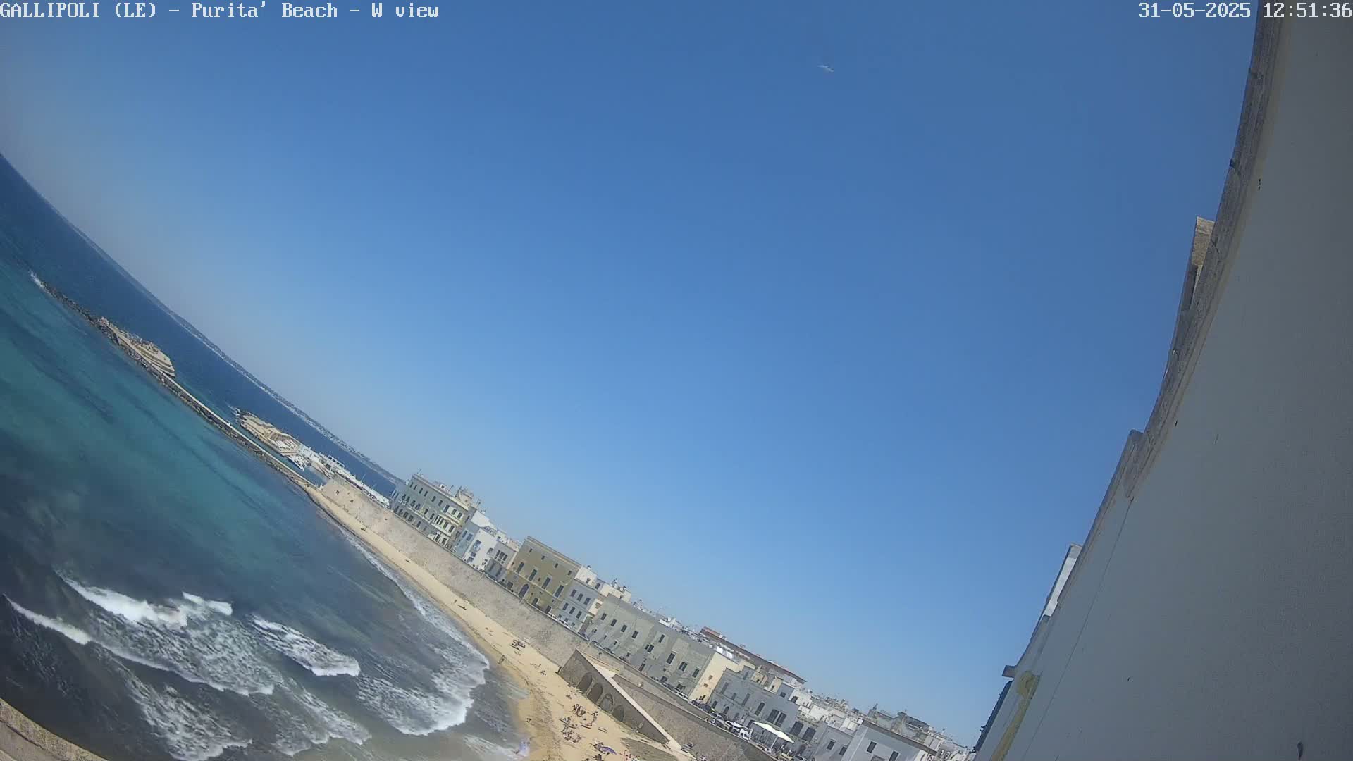 A beach with turquoise water and small waves laps against a sandy shore, next to a line of buildings under a clear blue sky.