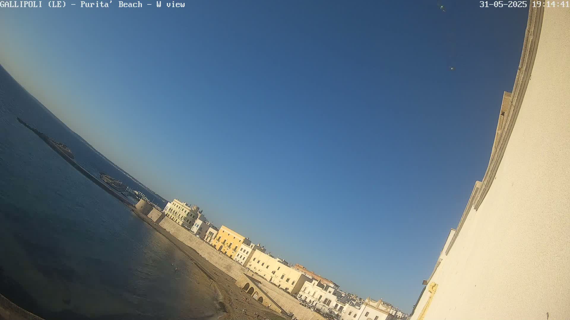 A clear blue sky overlooks a sandy beach and a row of buildings along a calm body of water.
