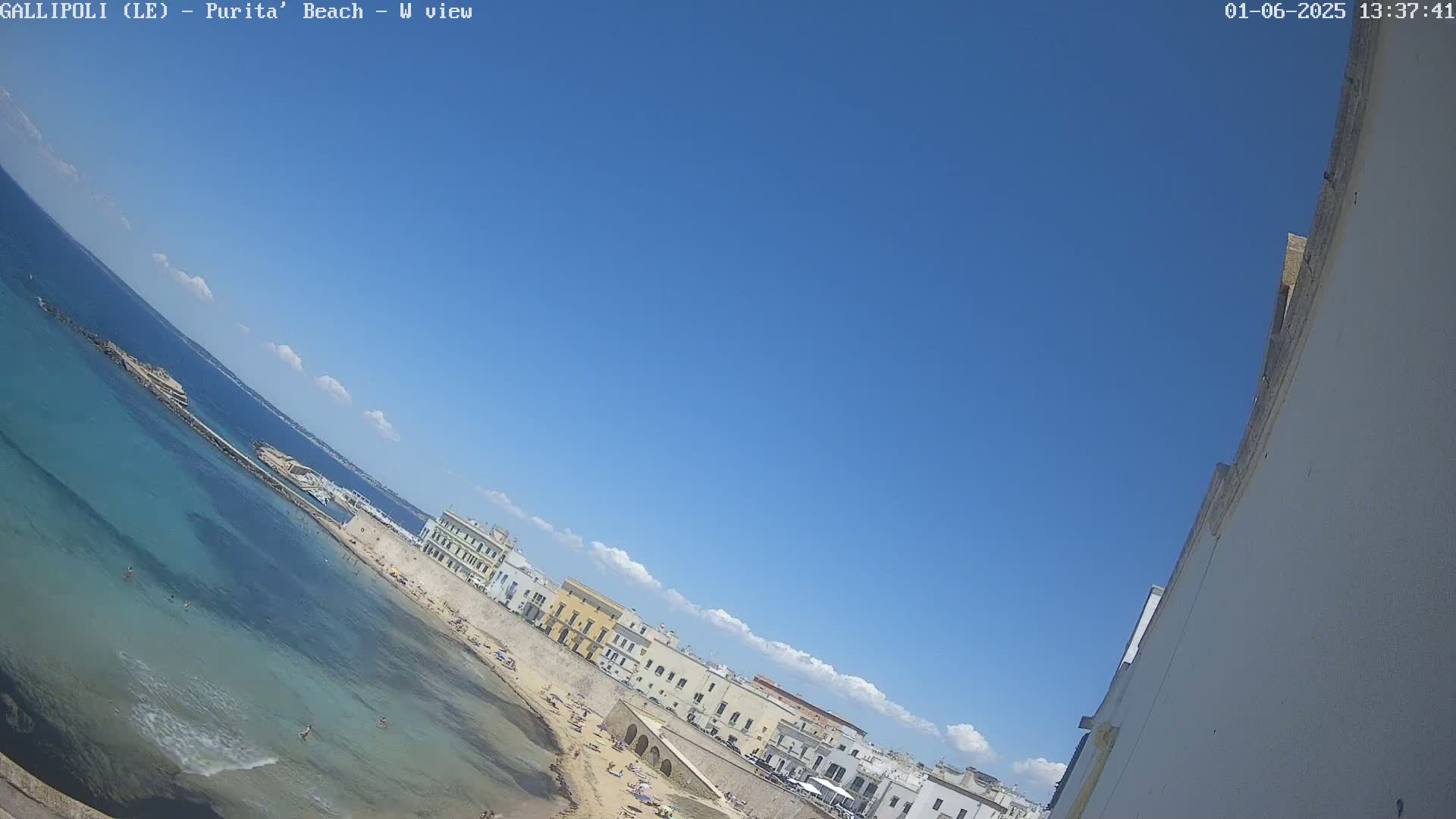 A mostly sunny day shows a beach with clear turquoise water and light-colored buildings along the shore.