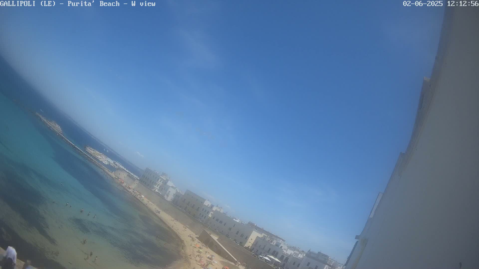 A clear blue sky overlooks a beach with light-colored buildings, clear shallow water, and people swimming and sunbathing.