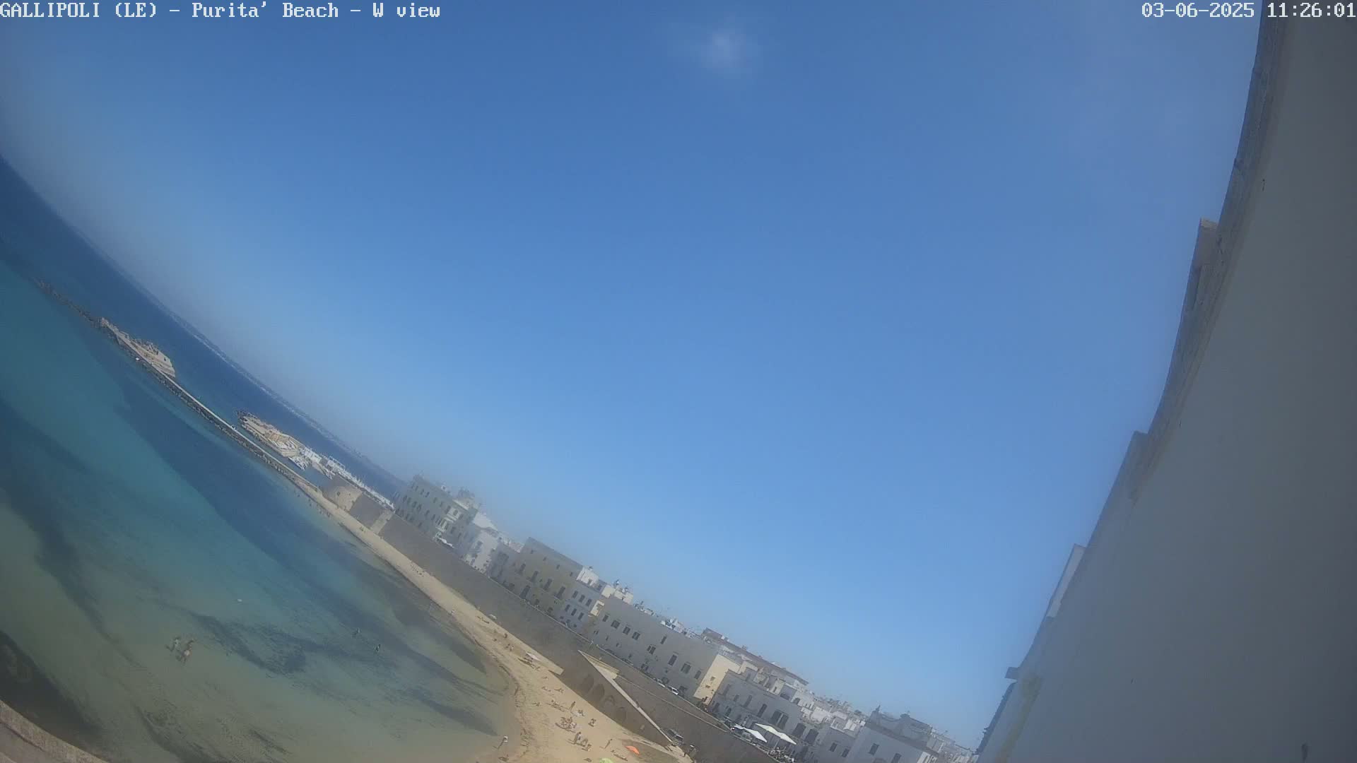 A clear, sunny day shows a beach with shallow, clear turquoise water next to a white building on a coastline.