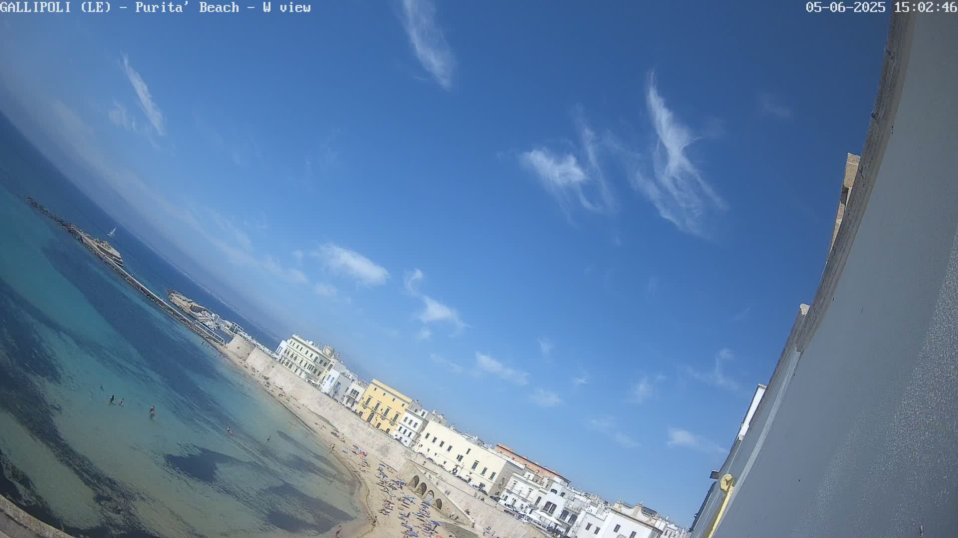A sunny day on a sandy beach with clear blue water, a row of buildings along the shore, and a few people swimming and sunbathing.