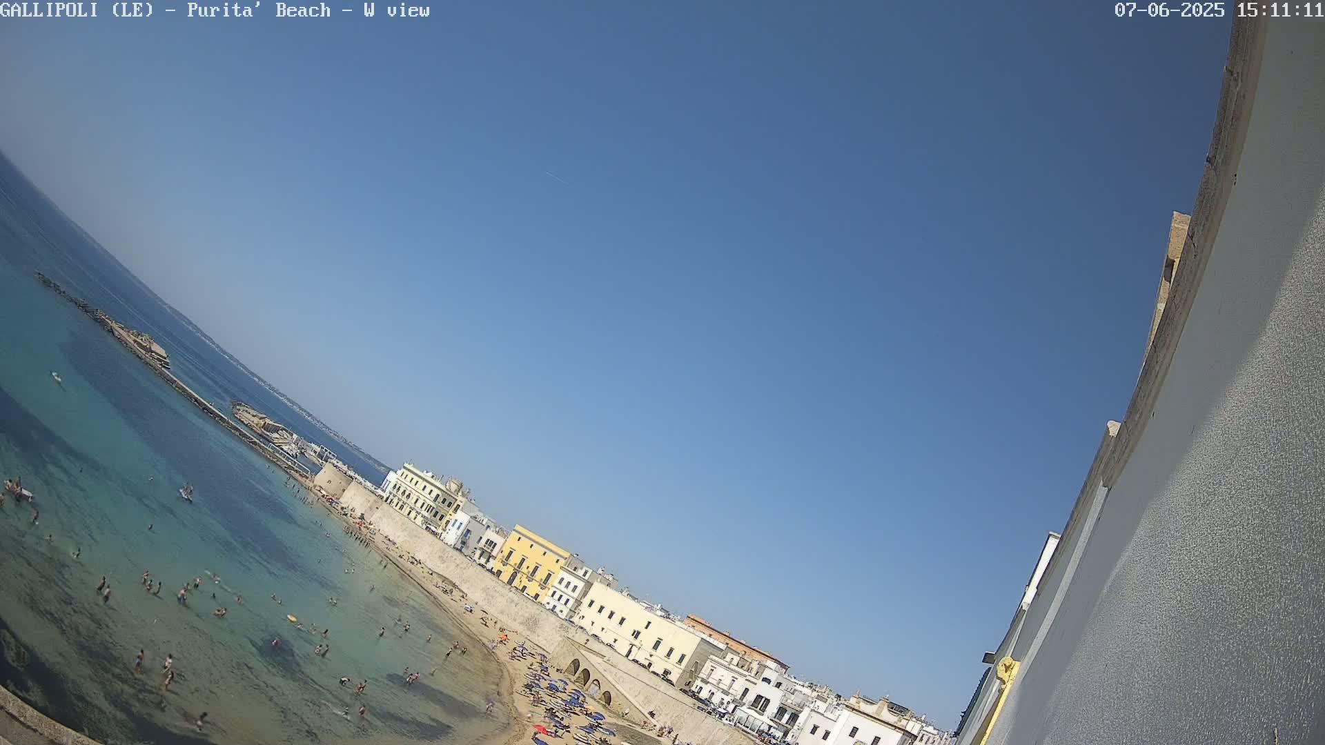 A clear blue sky overlooks a beach scene with many people swimming and sunbathing, adjacent to colorful buildings along a stone wall.