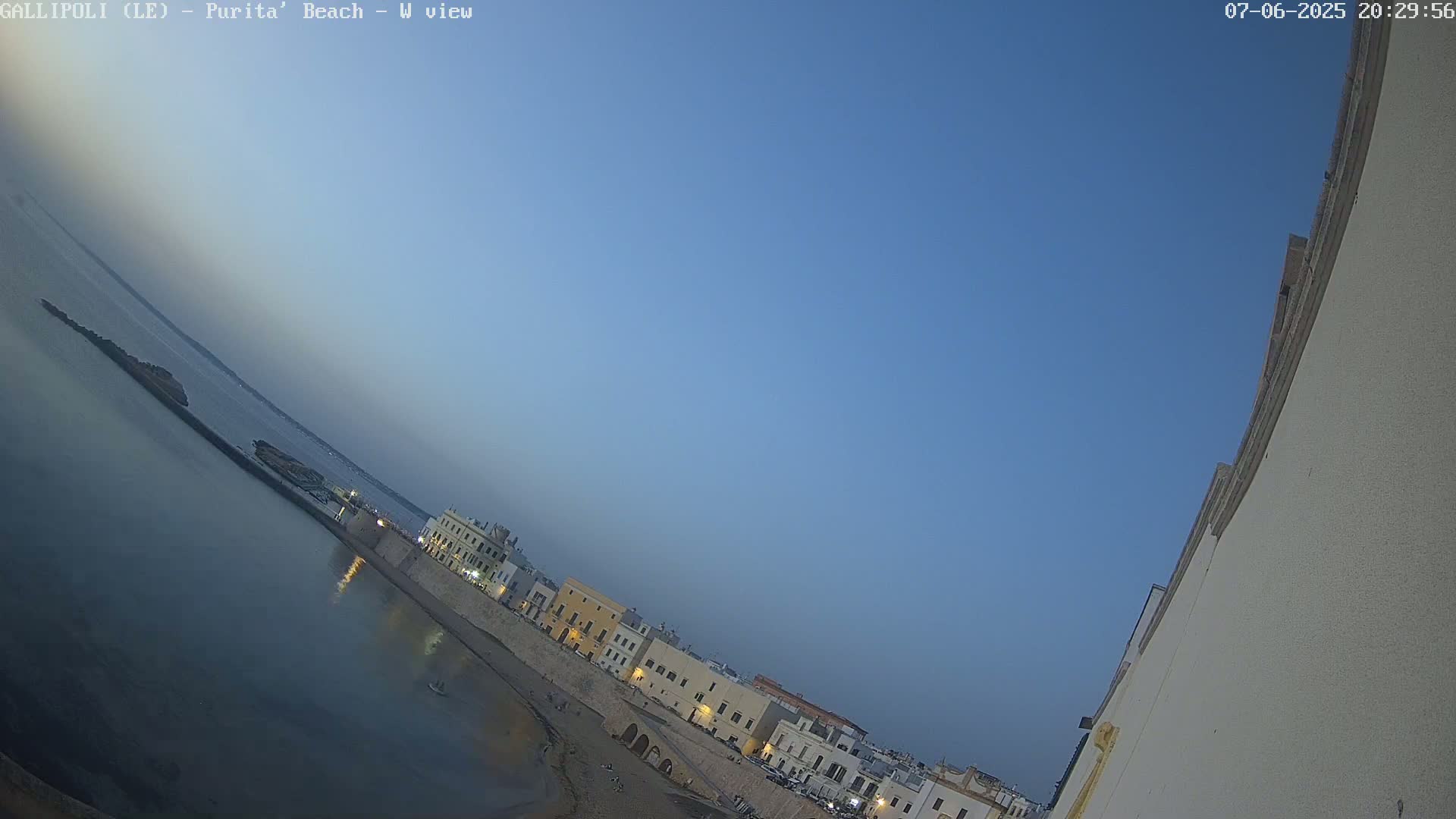 A seaside town at twilight, under a clear blue sky, is viewed from a high vantage point, showing a calm sea and buildings along a sandy beach.