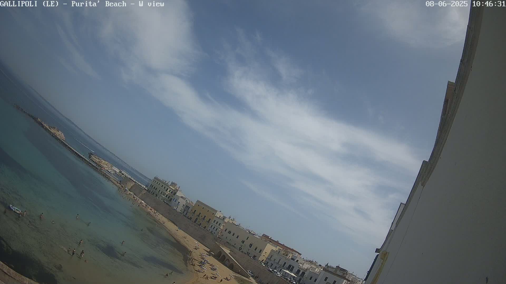 A sandy beach with clear, shallow turquoise water and several people swimming and sunbathing is next to a walled town under a mostly sunny sky with wispy clouds.