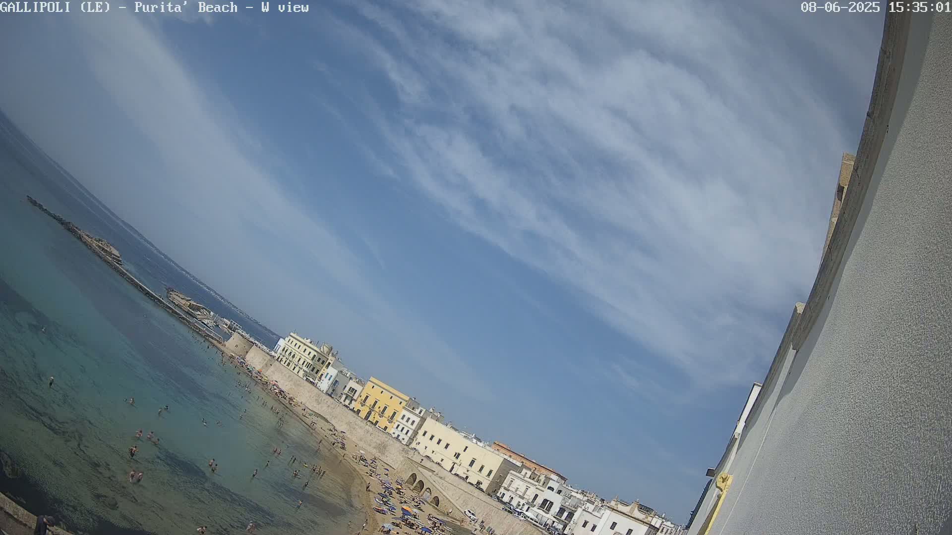A partly cloudy blue sky overlooks a sandy beach with clear, shallow water and many people swimming and sunbathing, adjacent to a line of buildings along a stone wall.