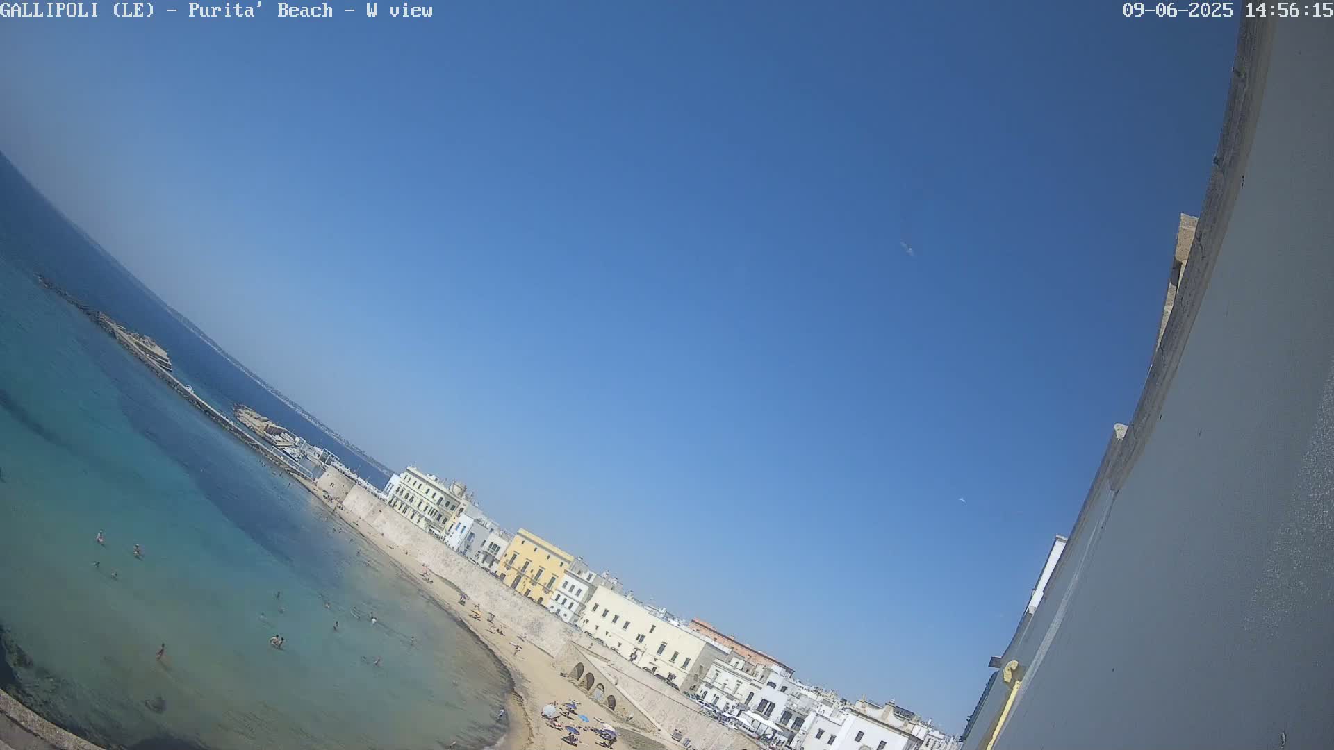 A sunny day shows a beach with clear turquoise water, people swimming and sunbathing on a sandy shore lined with buildings along a stone wall.