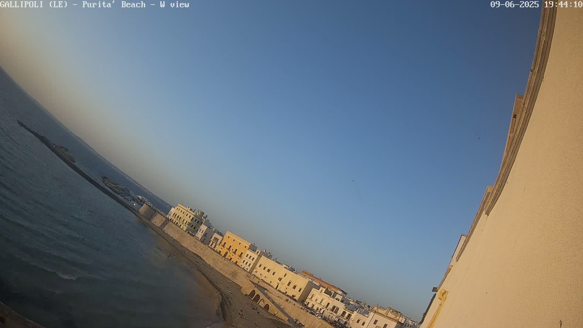 A beach with calm water and a row of buildings along a wall under a clear blue sky.