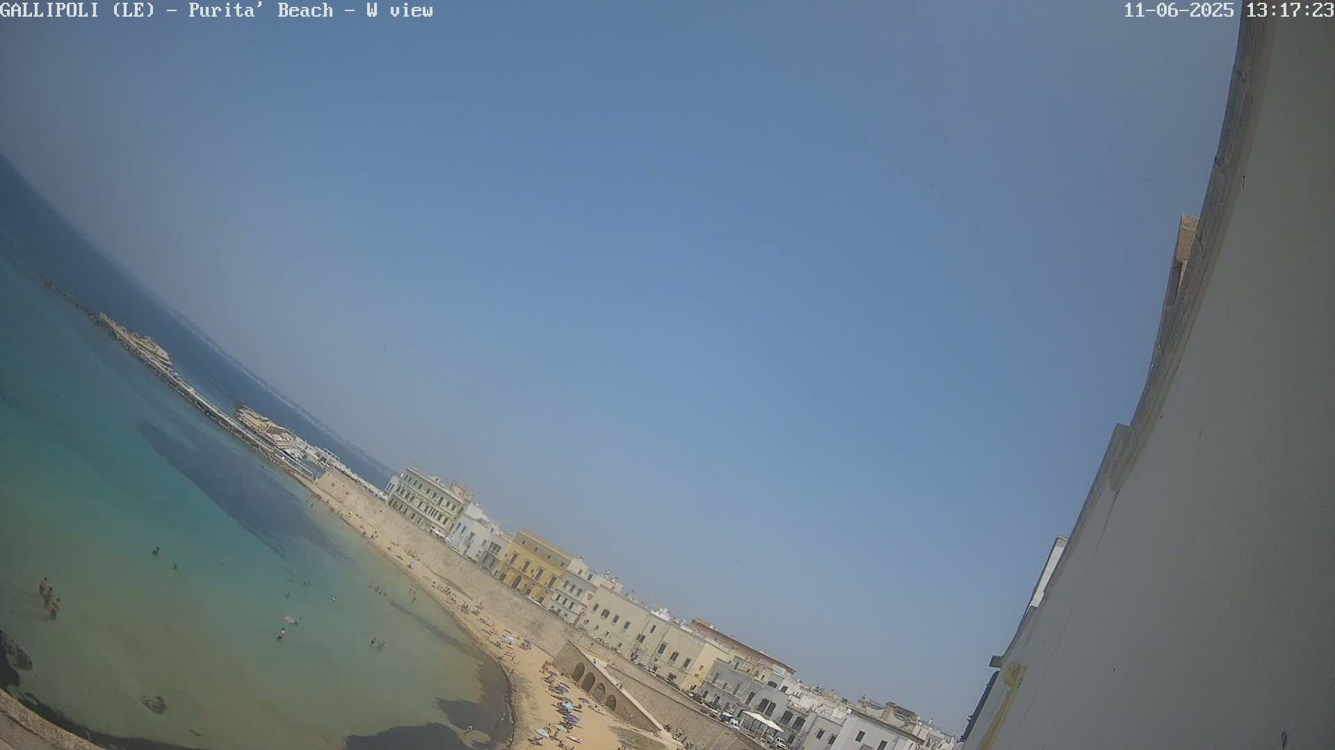 A clear blue sky overlooks a sandy beach with shallow, clear turquoise water and several buildings along the shoreline.