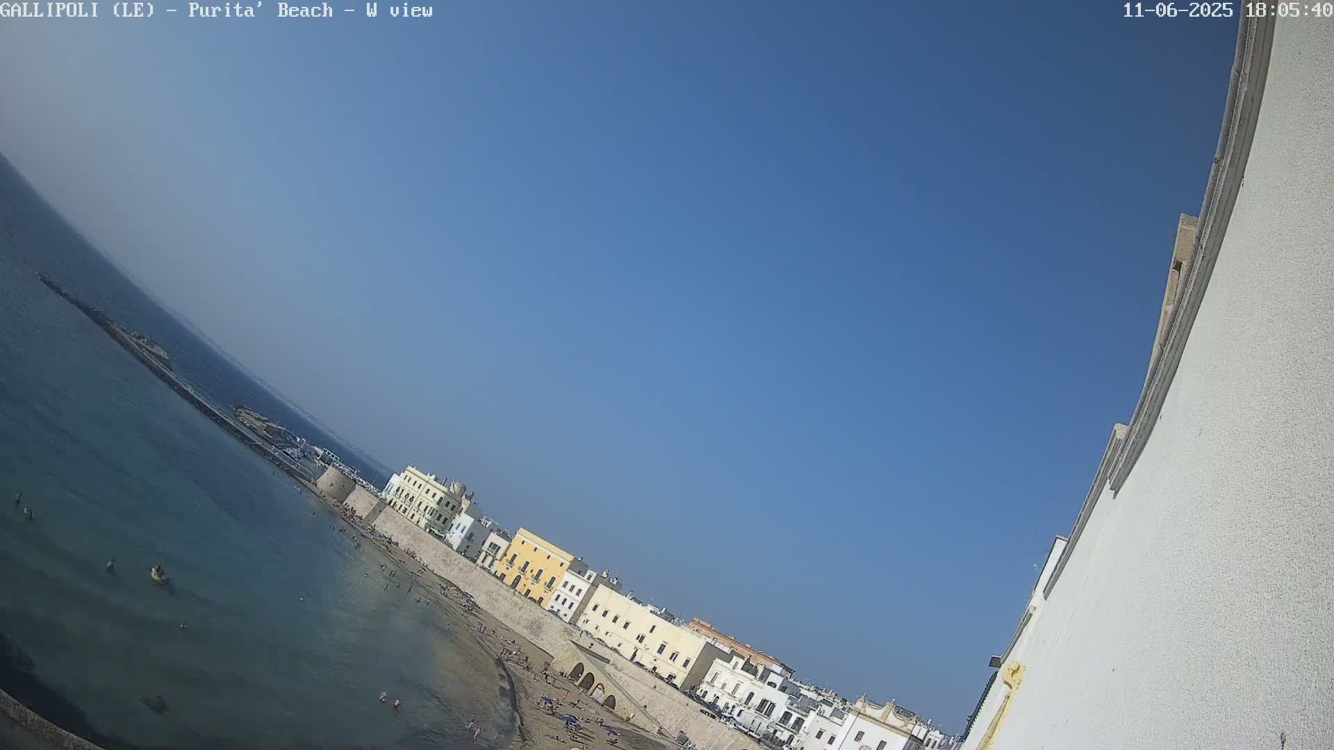 A sandy beach with clear blue water and buildings along the shore under a clear blue sky.