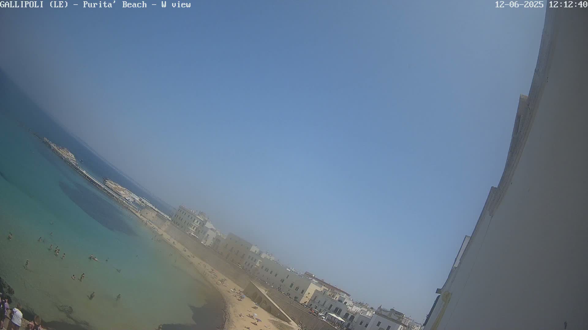 A clear, sunny day shows a beach with light-colored sand, clear turquoise water with people swimming, and buildings lining the shore.