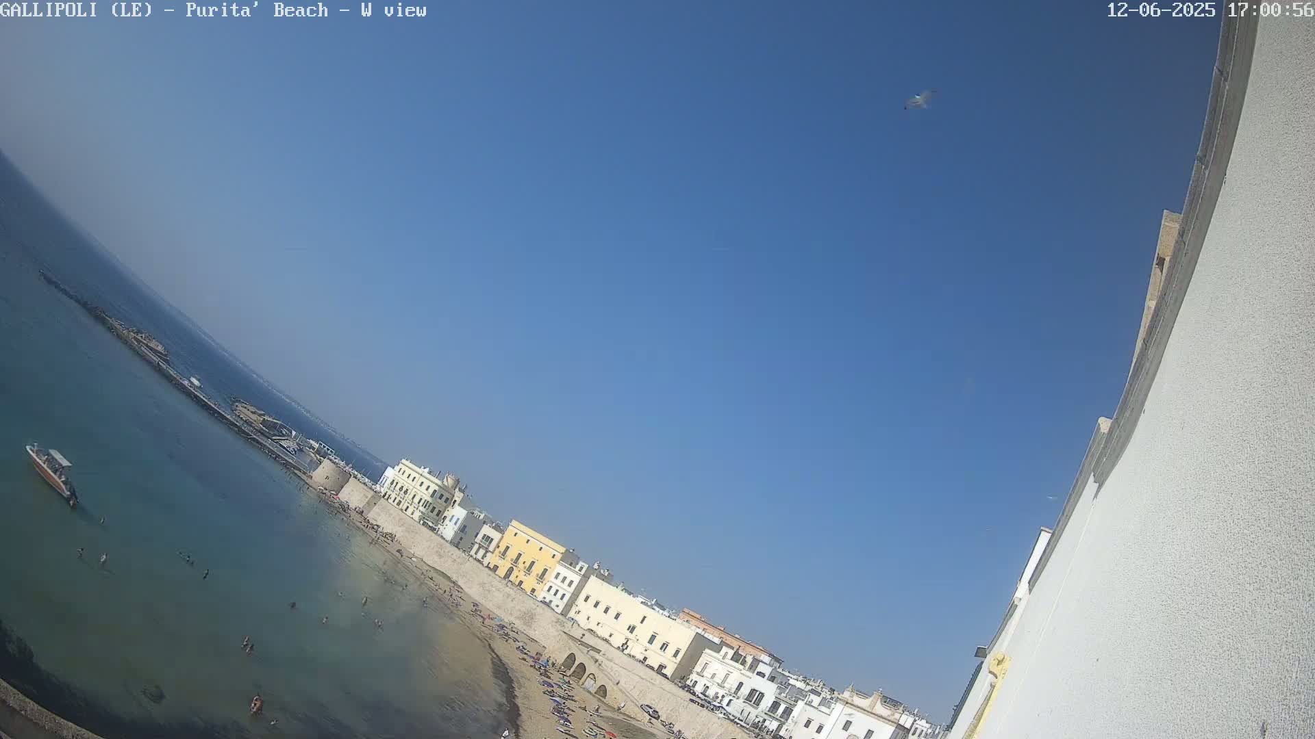 A beach with clear, shallow turquoise water, a sandy shore lined with people, and a row of buildings under a clear blue sky.