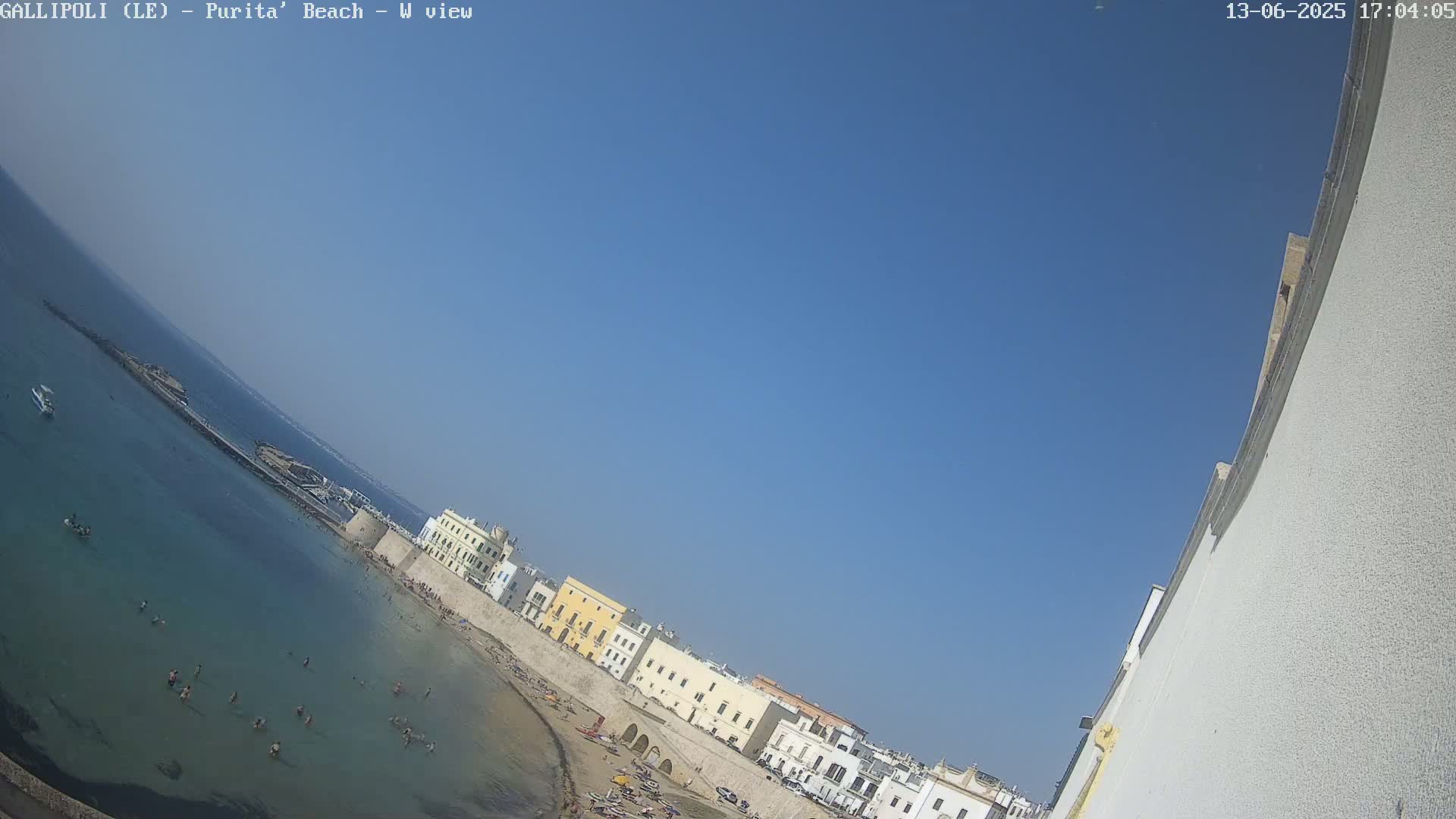 A clear blue sky is over a sandy beach with calm, clear water where people are swimming and sunbathing in front of a row of colorful buildings along a stone wall.