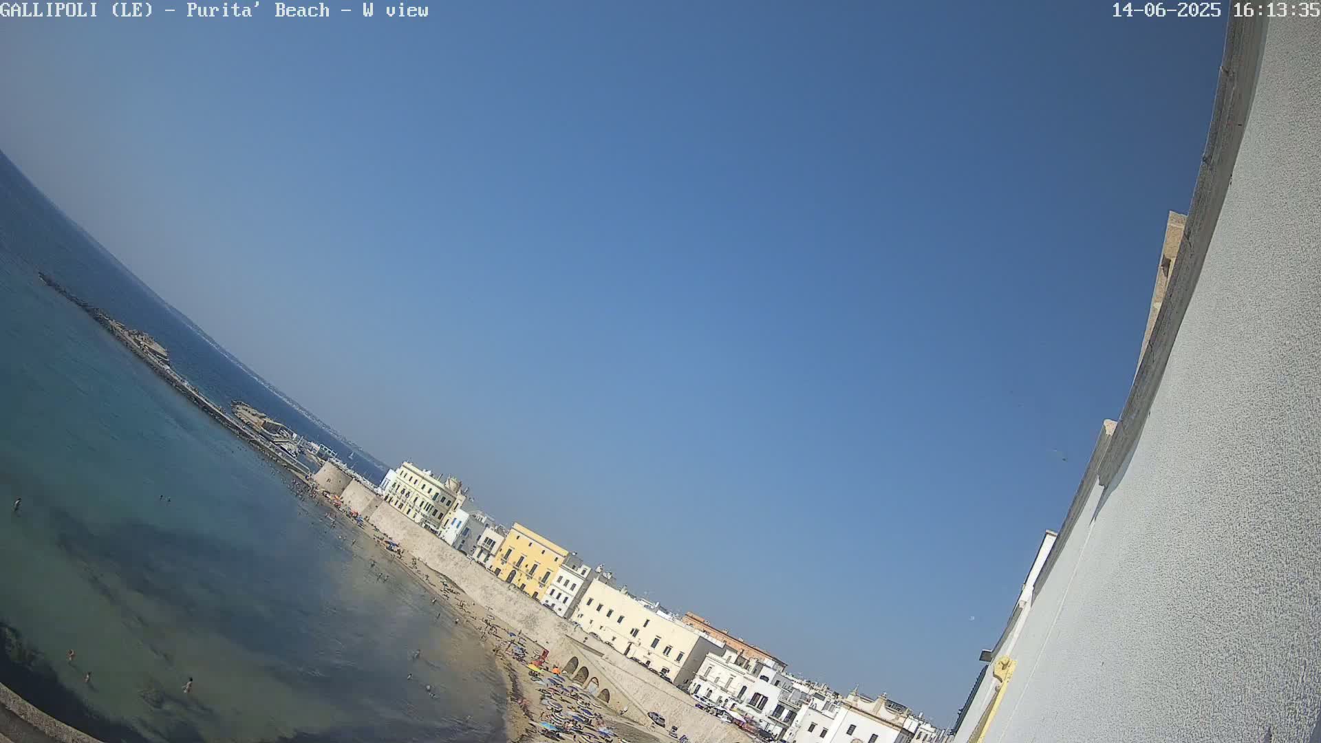 A clear, sunny day shows a beach scene with calm, clear water, a sandy shore with people, and buildings along the waterfront.