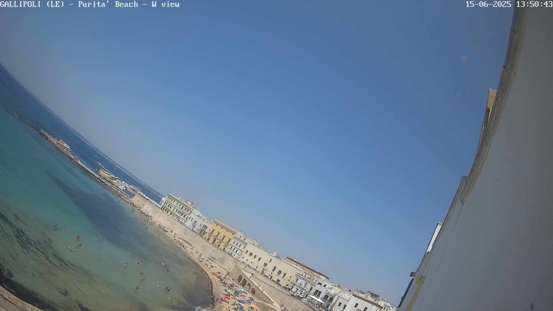 A beach with clear, shallow turquoise water and a sandy shore lined with low buildings under a clear blue sky.