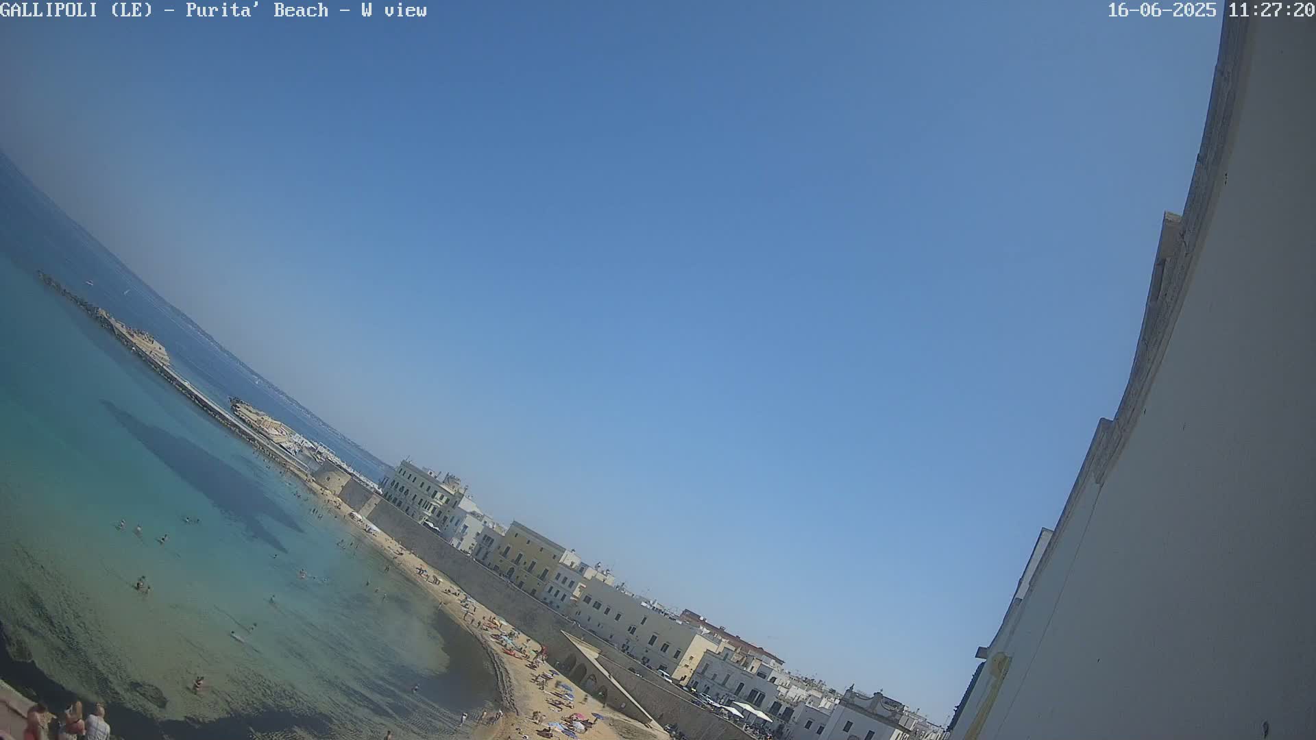 A clear blue sky overlooks a sandy beach with clear turquoise water, where people are swimming and sunbathing, next to a stone wall and buildings.
